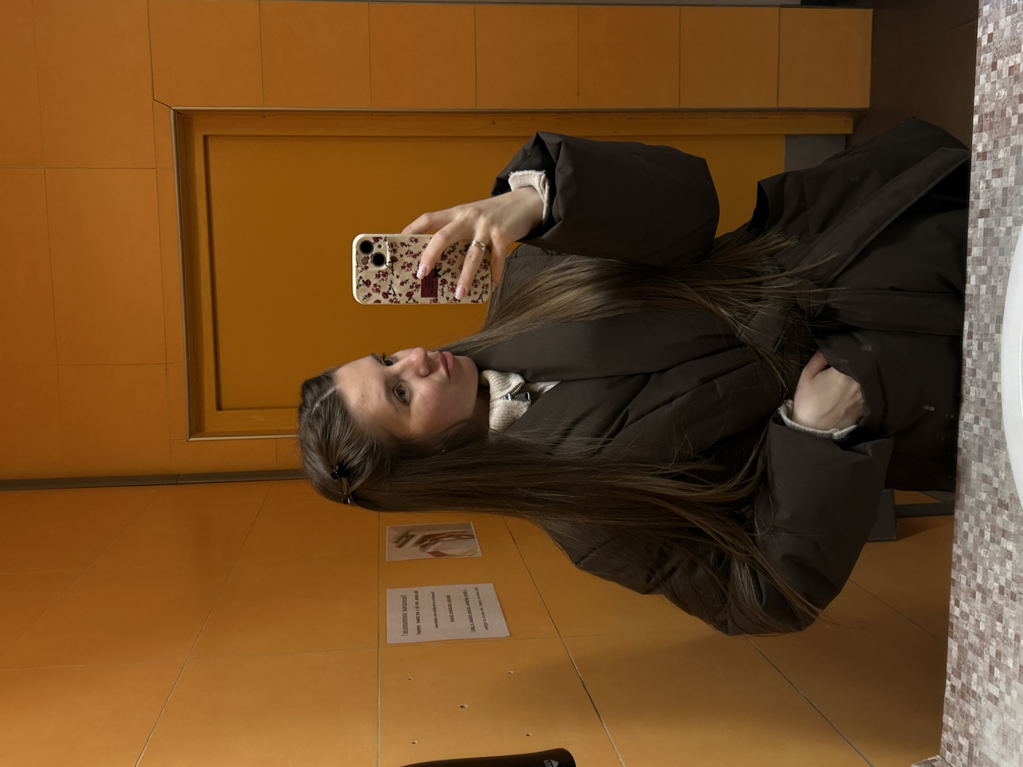 The image shows a woman in a bathroom. She is wearing a brown puffer jacket over a white collared shirt. She is holding a cell phone with a floral case in her right hand, taking a selfie in the mirror. The bathroom has orange tiled walls and a light-colored tiled countertop with a white sink. There are informational signs on the wall. Based on the text visible on the sign, the location is the city of Klin, Russia.