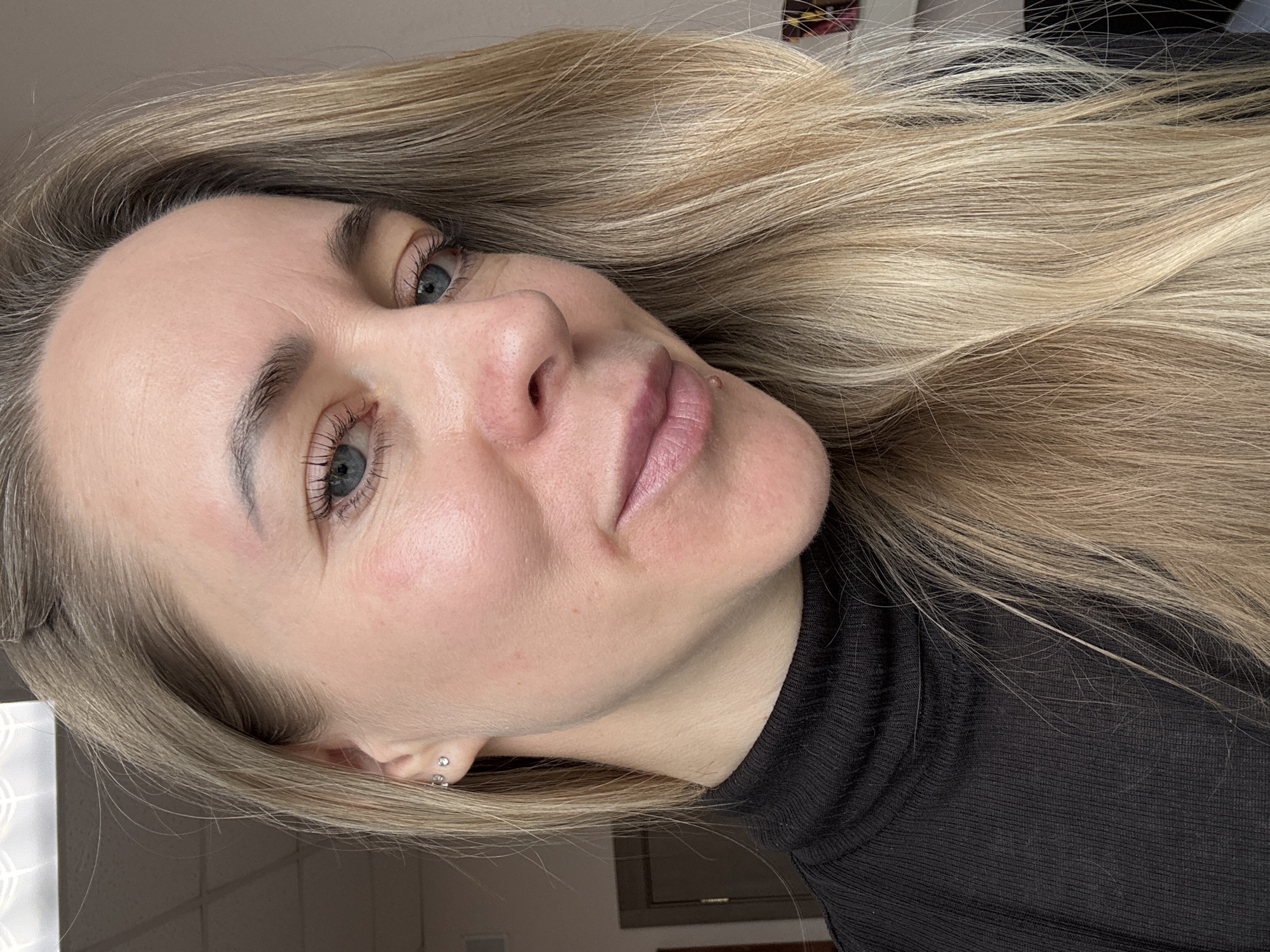 A close-up portrait shows a woman with long, flowing blonde hair, positioned horizontally, but the woman's head is tilted upwards and to the right, suggesting she is reclining or leaning back. She has bright blue eyes, with visible eyelashes, and naturally defined dark eyebrows. Her skin is fair, with a slight flush on her cheeks and some faint lines visible on her forehead. A small, light-colored blemish or mole is visible near her right nostril, above her upper lip. Her lips are a natural light pink. She is wearing a black, ribbed turtleneck top. In her visible left ear, she has small, silver-toned stud earrings. The background is indistinct, appearing to be an indoor setting with muted colors and some soft lighting. There are no specific distinguishing features in the background, though faint shapes suggest a room interior. The woman's expression is neutral and calm. There is no visible text in the image. The location context provided is Chaikovsky, Russia.
