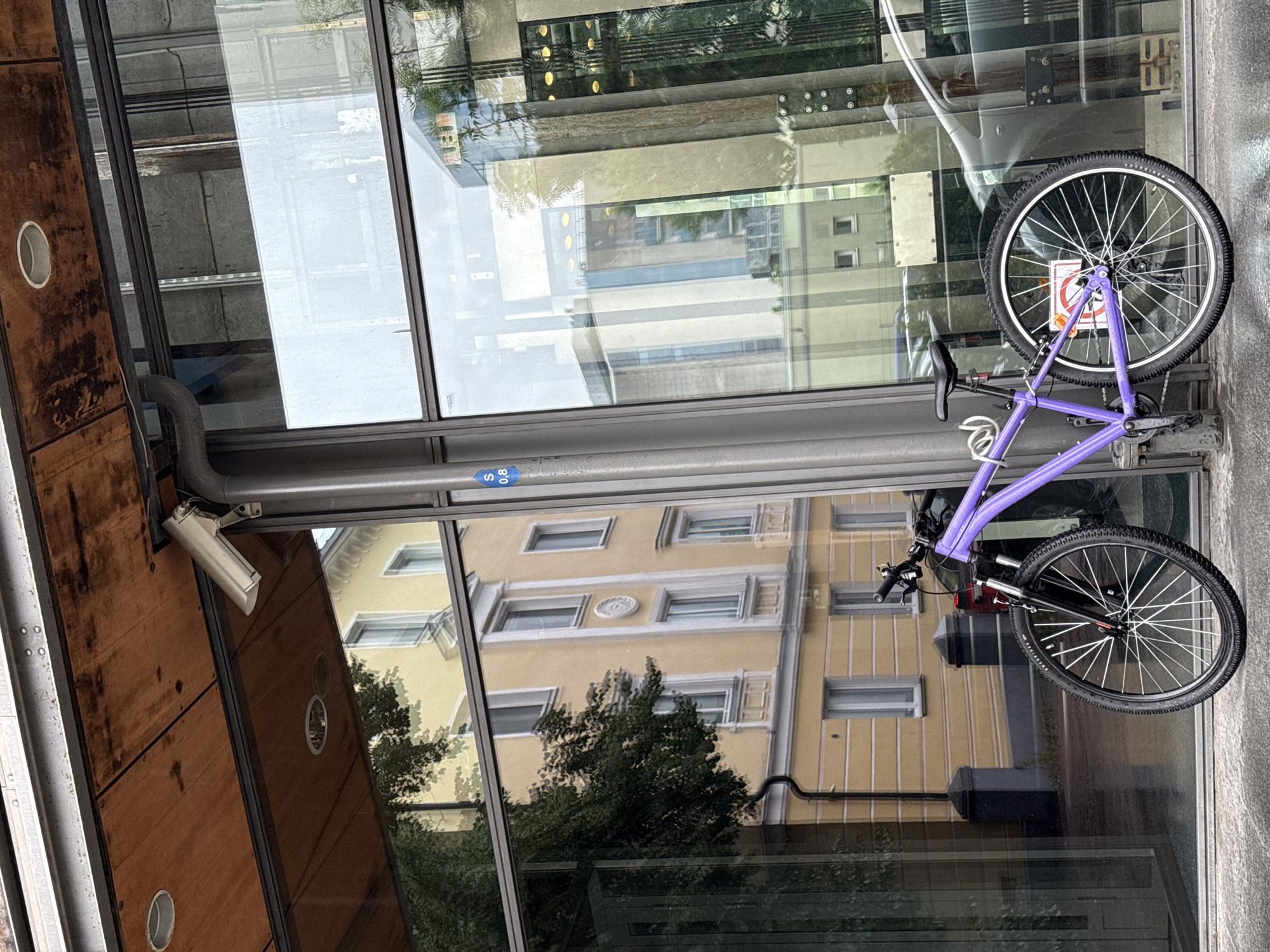 A purple bicycle is parked next to a modern building with large glass windows. The bicycle has a black seat, black handlebars with a bell, and black wheels with silver spokes. A silver lock is attached to the frame. On the rear wheel, a sign with a red circle and a diagonal line through a cigarette is visible.
The building's exterior features a wooden overhang with a security camera mounted beneath it. The glass windows reflect buildings across the street, including a yellow building with several windows and decorative white trim. A blue sticker with "S 0,8" is affixed to a metal post near the bicycle. The ground appears wet, suggesting it may have recently rained. The scene is set in Tallinn, Estonia, likely in a business or commercial district.