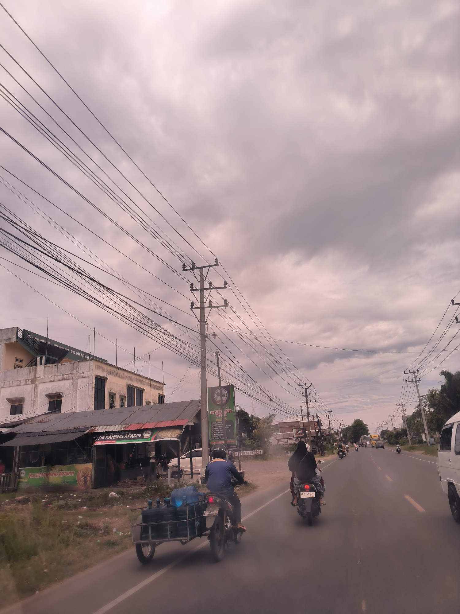 The image presents an active street scene in Payameuneng, Indonesia, captured on an overcast day with a sky full of grey clouds. The perspective appears to be from a vehicle moving along a paved road, looking forward.
In the foreground, a man on a motorcycle is seen from behind, pulling a sidecar laden with several large blue water jugs. To his right, another motorcycle carries two people, with the passenger wearing a dark head covering. Further down the road, additional motorcycles and a white van are visible, suggesting moderate traffic.
Along the left side of the road, there are several buildings, including a multi-story structure and a line of smaller, open-fronted shops or stalls with corrugated metal roofs. Prominent signs above these shops display text such as "BU SIE KAMENG APADIN" and "BUSIEK MENG APA IN," indicating local businesses, possibly eateries. Numerous dense power lines crisscross the sky, supported by a series of utility poles that line both sides of the street, dominating the upper portion of the frame. Visible license plate numbers include "BL 6739 ZAT" on one of the motorcycles and a partial "BG5501日" on another.
The overall scene depicts a slice of daily life, characterized by local commerce, typical street infrastructure, and common modes of transport in a busy urban or semi-urban setting.