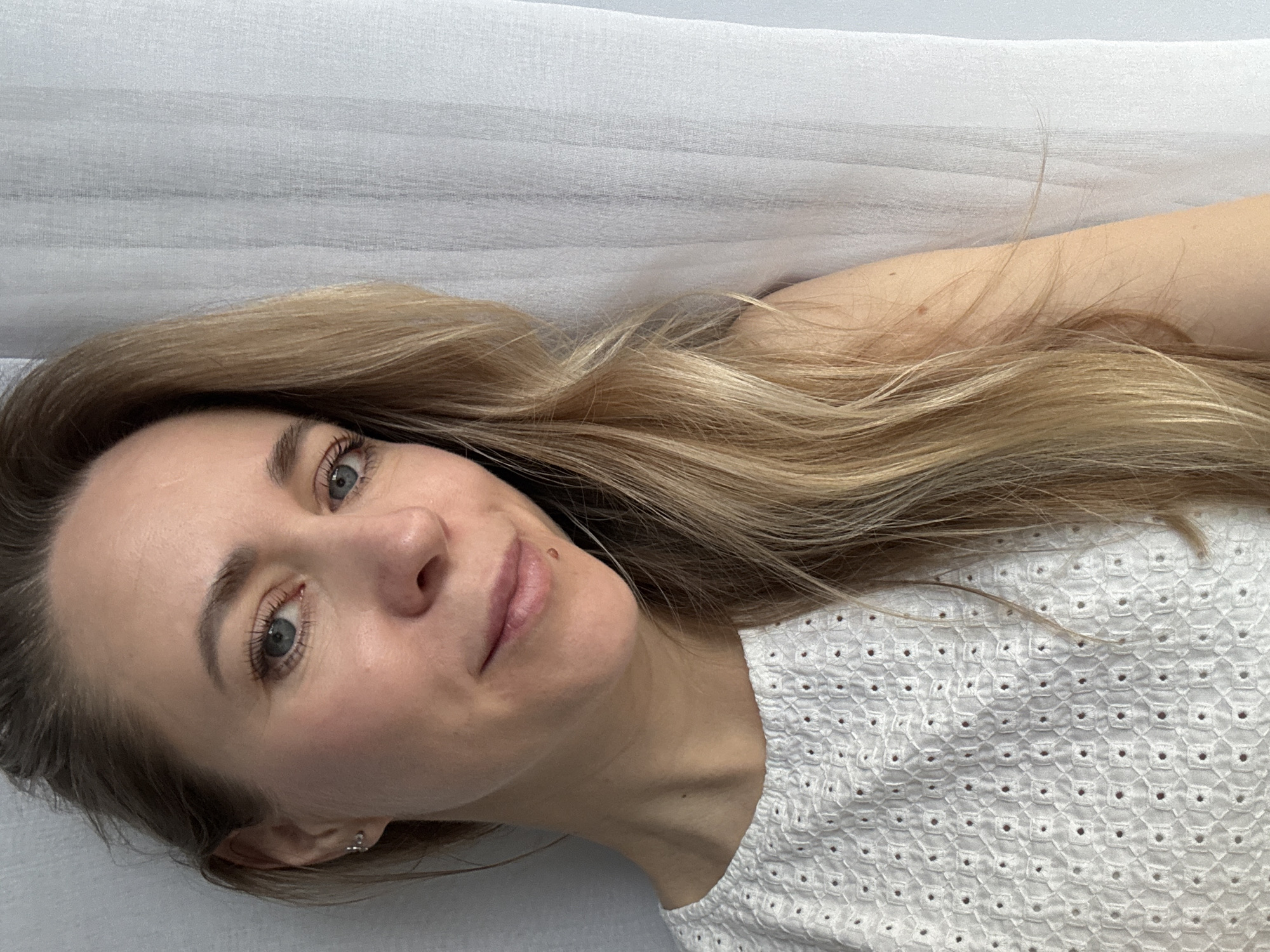This rotated portrait, taken in Chaikovsky, Russia, features a woman in her 30s or 40s lying down and looking directly at the camera with a calm, content expression. Her head is positioned towards the left of the frame, with her body extending to the right. She has long, flowing blonde hair, fair skin, and striking blue-green eyes. A small mole is visible above the right side of her upper lip.
She is wearing a white top with a distinctive eyelet embroidery pattern, composed of small, square-like cut-outs arranged geometrically. A subtle earring can be seen in her visible left ear. The setting appears to be indoors, against a light-colored, possibly white or very light grey, textured fabric background, which could be a pillow, bedsheet, or curtain, displaying faint horizontal lines or creases. The lighting is soft and even, indicative of indoor ambient conditions. There is no visible text in the image.