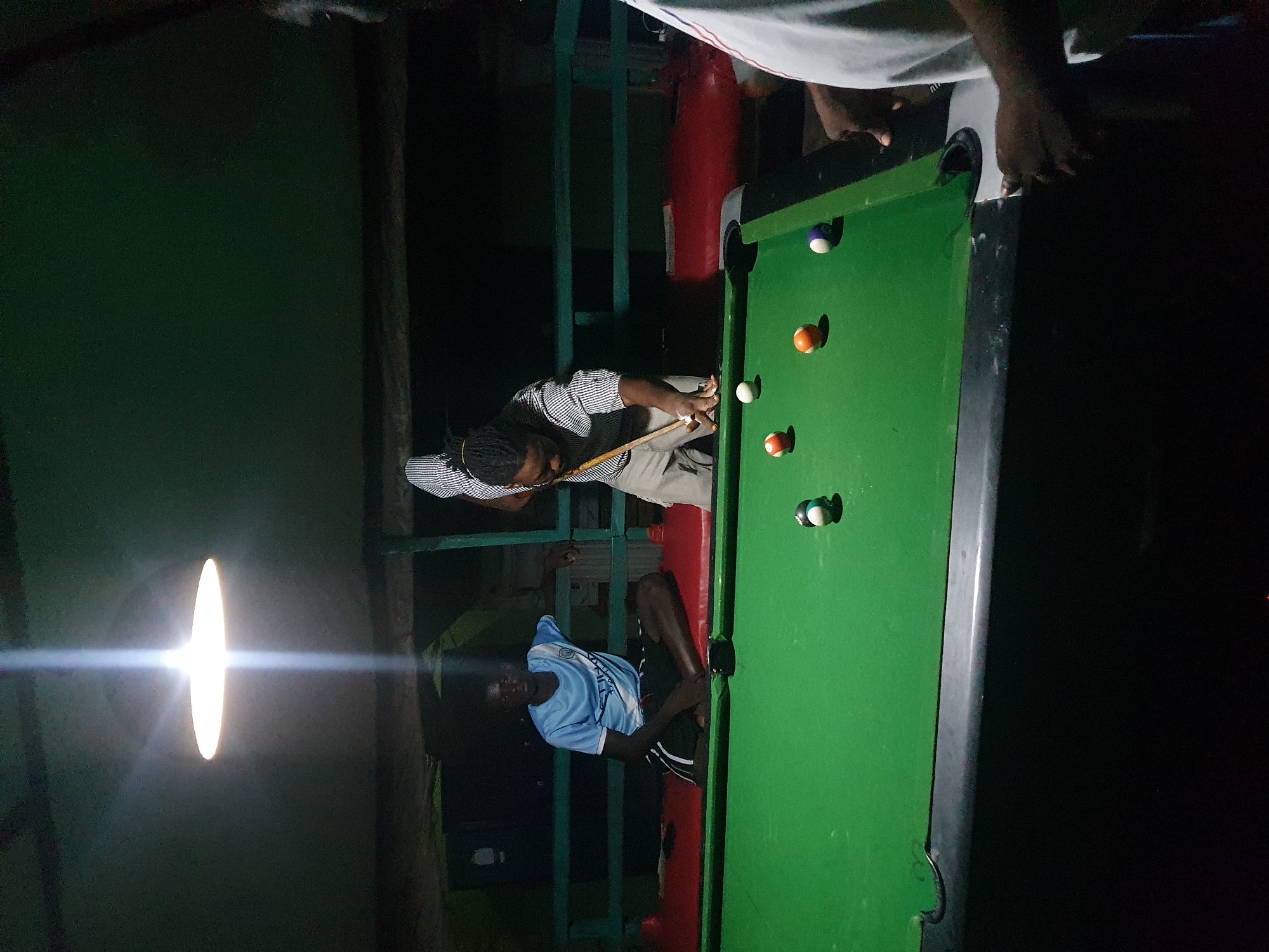 The image depicts a dimly lit indoor pool hall, likely in Lokoja, Nigeria. A man with dreadlocks, wearing a striped shirt and light-colored pants, is actively playing billiards, aiming a cue stick at a ball on the green felt table. Another man, wearing a light blue t-shirt and dark shorts, is seated on a red bench to the left, observing the game. Several billiard balls, including a white cue ball, orange and white striped balls, and a blue and white ball, are scattered on the table. A bright overhead light fixture illuminates the playing area. The environment appears somewhat rustic, with a dark ceiling and green metal railings visible in the background. The focus is on the game of billiards and the two individuals engaged in or observing it.