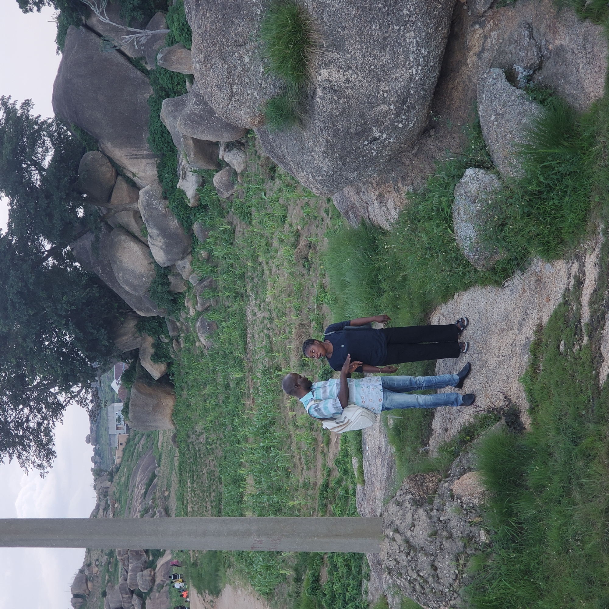 A man and a woman are standing on a dirt path in a rocky, vegetated area. The man is wearing a blue and white striped shirt, blue jeans, and black shoes. He has a white backpack slung over his left shoulder and is holding a phone in his right hand, looking down at it. The woman is wearing a dark blue t-shirt, black pants, and sandals. She is looking at the man and appears to be speaking to him, with her mouth open. The path is surrounded by sparse green vegetation and large grey rocks. In the background, there is a field of crops, more rocky terrain, and a few buildings. This scene appears to be in the city of Jos, Nigeria, a region known for its rocky landscapes and agricultural activity.