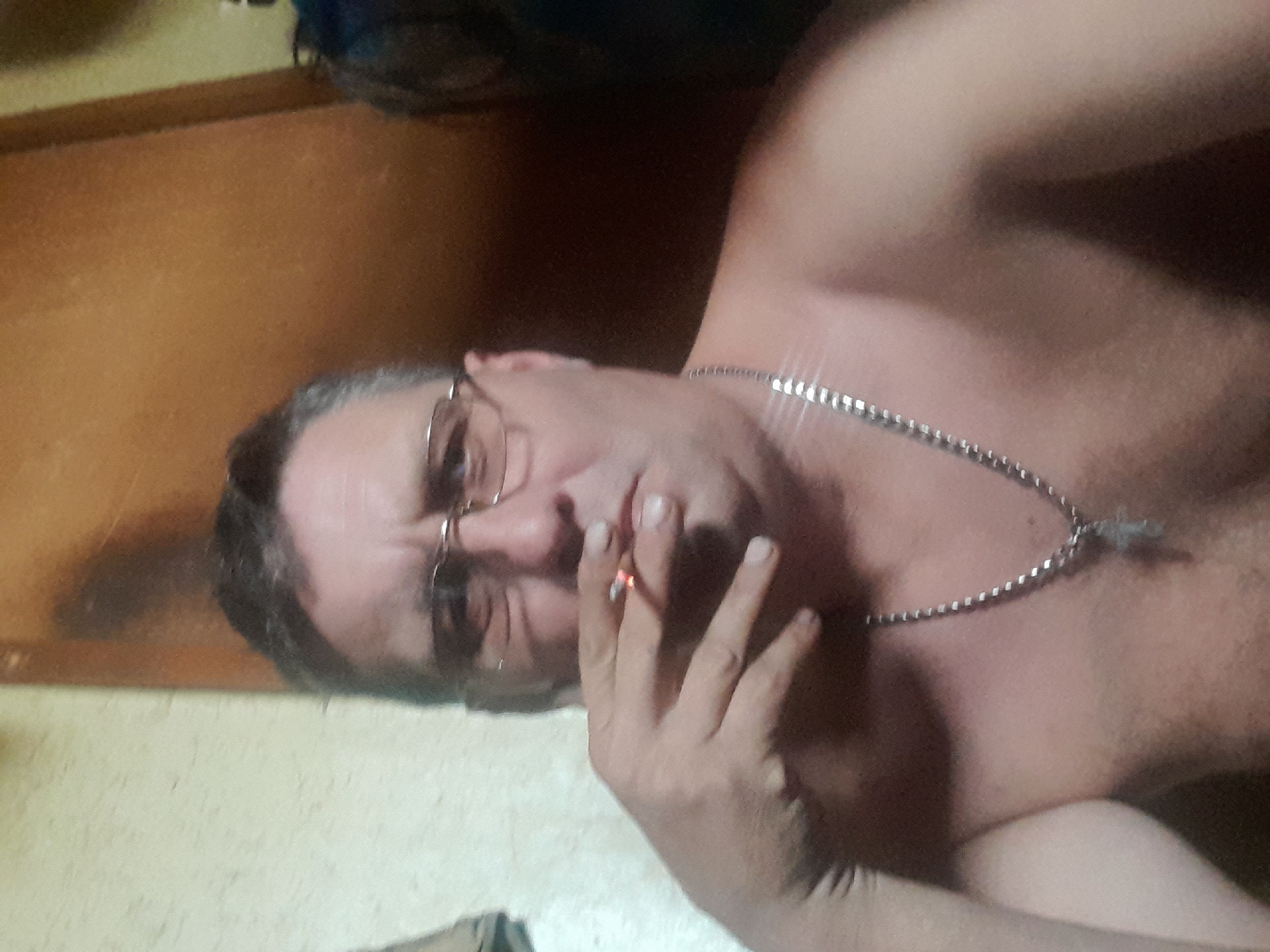 A shirtless man with glasses is holding a lit cigarette to his lips. He is wearing a silver chain around his neck. The background appears to be indoors with wooden paneling. No specific location in Volokonovka, Russia can be determined from the image.