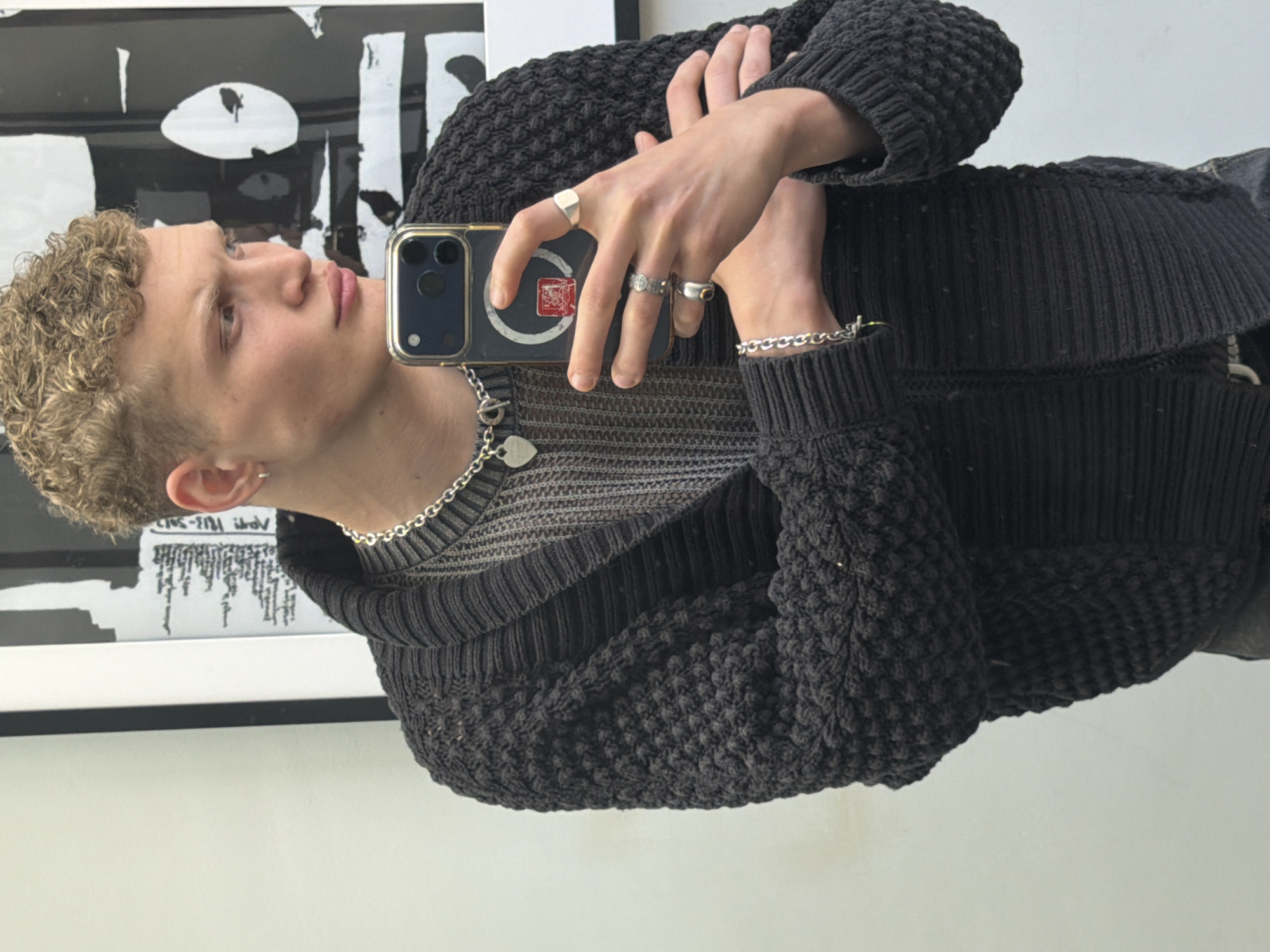 A young, light-skinned man is taking a selfie in front of what appears to be a piece of art. The art has a black and white design. The man has short, curly blonde hair, and he is wearing a silver chain necklace with a heart pendant, and a silver bracelet. He is also wearing multiple silver rings on his fingers. He is holding a smartphone, possibly an iPhone, with a blue case and a red and white sticker on the back. He is dressed in a dark grey sweater, and a dark knit cardigan. The location appears to be indoors, likely in London, United Kingdom, considering the style of the art and clothing are common in that area. The overall lighting is soft and diffused.