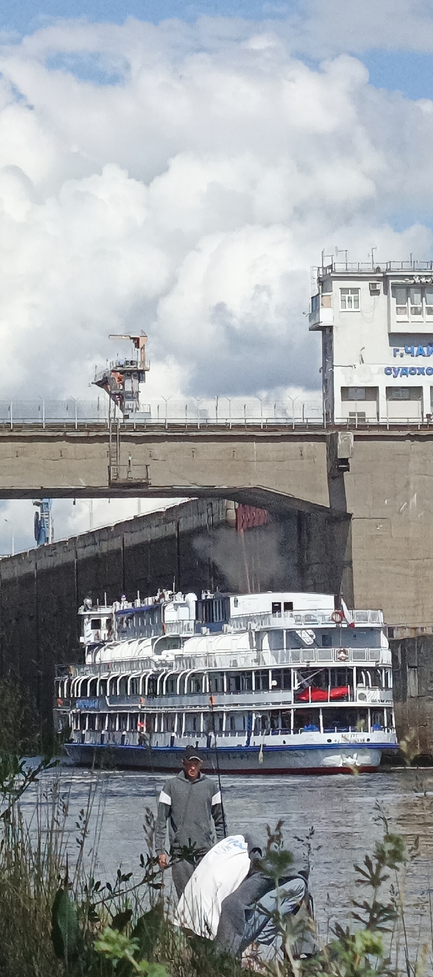 The image depicts a river lock with a multi-level river cruise ship positioned within it. The lock is constructed of concrete and features a control tower on either side, connected by a bridge. The name of the structure, starting with Г.ЧАЙ, is visible on one of the towers.
In the foreground, a person is seen fishing from the riverbank among vegetation. The sky is partly cloudy with patches of blue.
The location is presumed to be in an unspecified city, in an unspecified region, due to the incomplete inscription on the tower.
