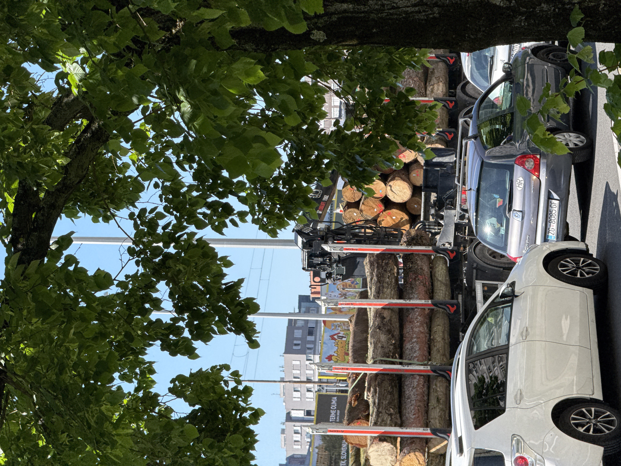 The image, rotated 90 degrees counter-clockwise, presents a bright daytime street scene, partially framed by the lush green leaves and sturdy trunk of a large tree in the foreground.
In the midground, a heavy-duty truck is prominently featured, loaded with numerous freshly-cut logs. The logs are neatly stacked horizontally, secured by distinctive red and white vertical supports, and many exhibit red paint markings on their cut ends. A crane arm, clearly labeled "LOGLIFT 1502," is visible on the truck, indicating its purpose for timber handling. Adjacent to the log truck, a dark grey Renault car is parked, displaying a Croatian license plate "ZG 8085-GF," which signifies Zagreb, along with an "HR" country sticker. Further to the right, a white SUV is also parked.
The background reveals several multi-story buildings under a clear blue sky, typical of an urban setting. Visible text on signs includes "TERME OLIMIA" and "SLOVENIJA," suggesting a potential connection to a spa resort in Slovenia or indicating the truck's route. A colorful billboard with cartoon characters and the word "Zmajček" is also discernible. The bright sunlight casts distinct shadows, confirming a sunny day. The scene captures a moment of commercial activity, specifically timber transport, within an urban environment in Zagreb, Croatia.