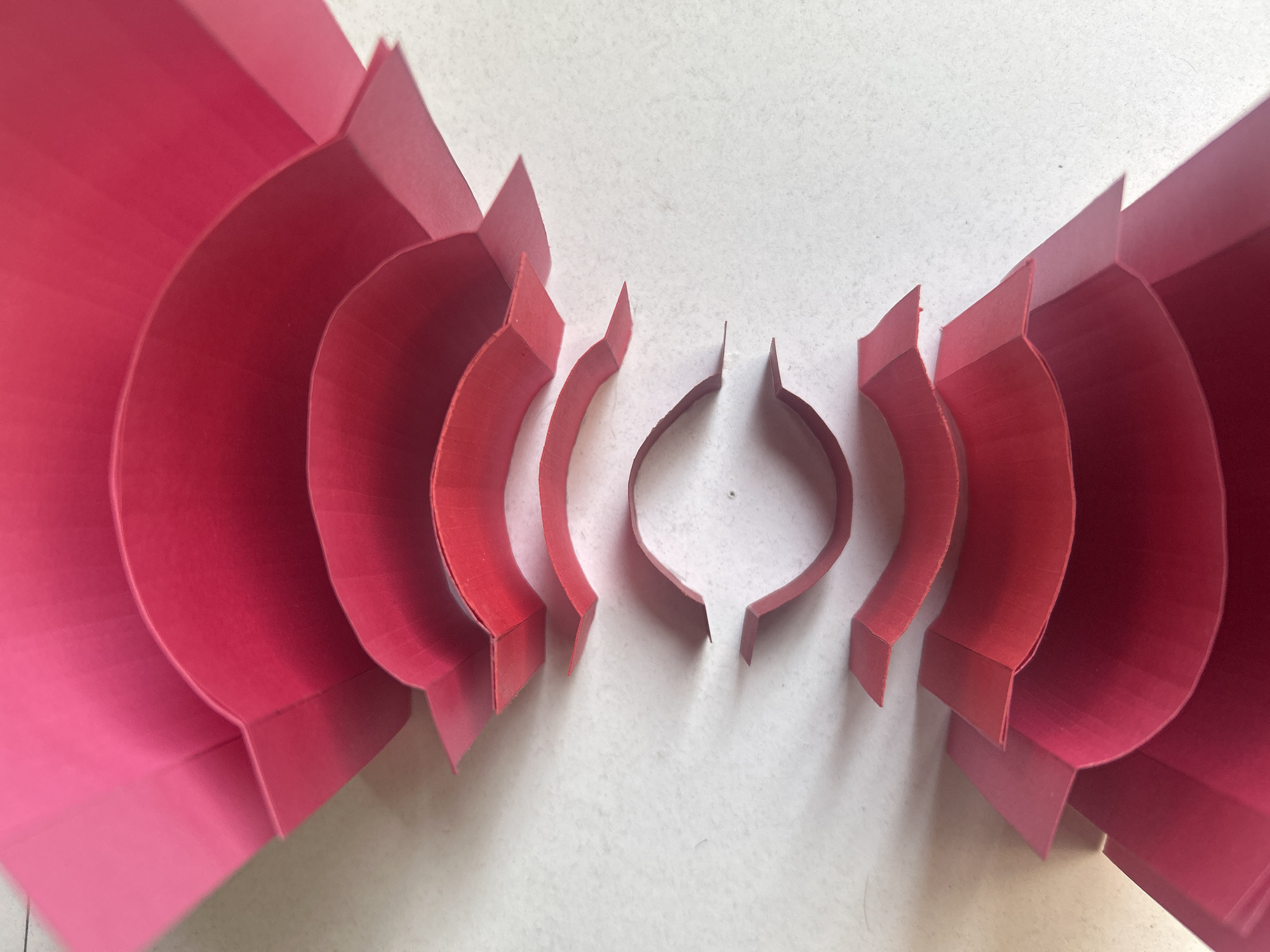 The image displays a symmetrical arrangement of red paper cutouts on a white, tiled surface. The cutouts are segmented arcs of increasing size, with each arc flanked by a perpendicular tab at its base. The arcs are arranged from smallest in the center to largest on the edges of the frame. The background is a white tiled surface.