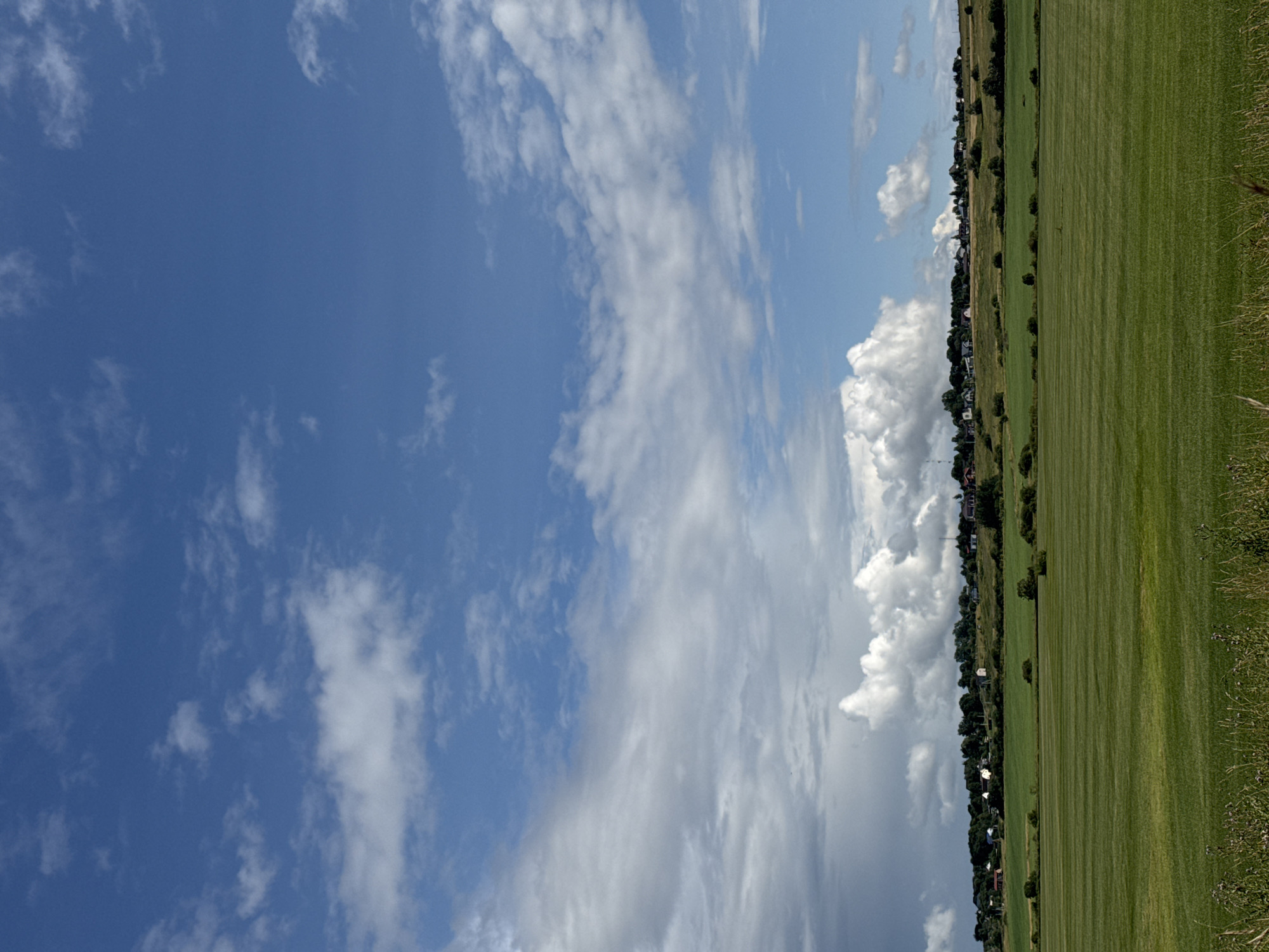 A wide, horizontal shot captures a rural landscape under a partly cloudy blue sky. In the foreground, a vast, well-maintained green field stretches across the frame, its surface marked by subtle horizontal lines suggesting it has been recently mowed or planted. Beyond this field, a gently sloping hill is dotted with trees and a collection of small buildings, indicative of a village. The buildings are varied in color and size, with some roofs visible. The sky is a dominant feature, filled with a mix of white, puffy cumulus clouds and some thinner, more wispy clouds, with patches of clear blue visible between them. To the left, the clouds appear darker and more dense, hinting at potential rain. The overall scene depicts a tranquil, agricultural environment in the village of Kempelevo, Russia. No people or animals are visible in the image.