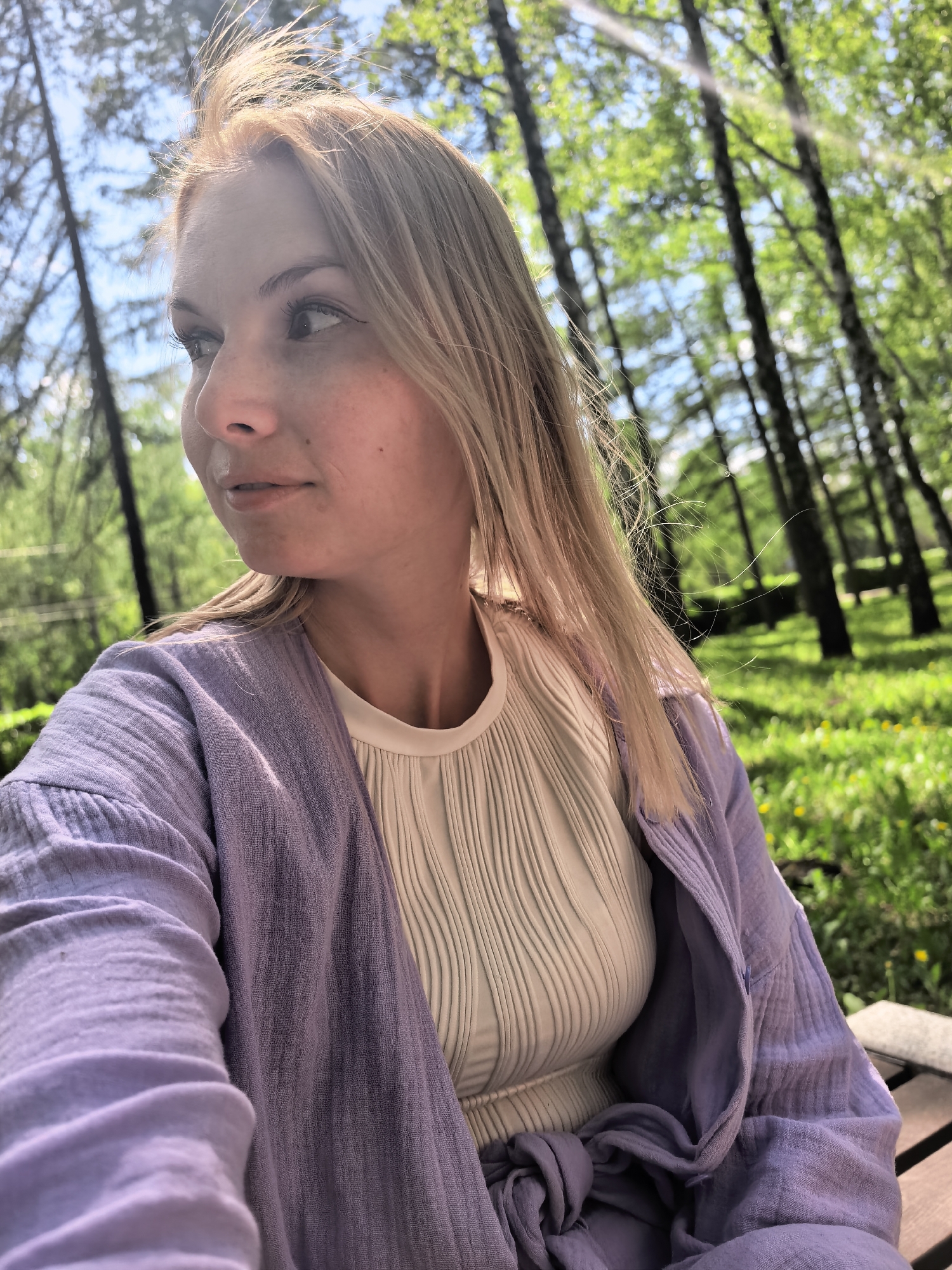 The image shows a woman with blonde hair, wearing a light purple cardigan over a white top, sitting outdoors in a park or wooded area. The background is filled with tall, thin trees with green leaves and a clear blue sky peeking through the branches. The location is in a green space of Sterlitamak, Russia. The woman is looking to the left, away from the camera. The lighting is bright, with sunlight filtering through the trees. The ground appears to be covered in grass and some yellow flowers.