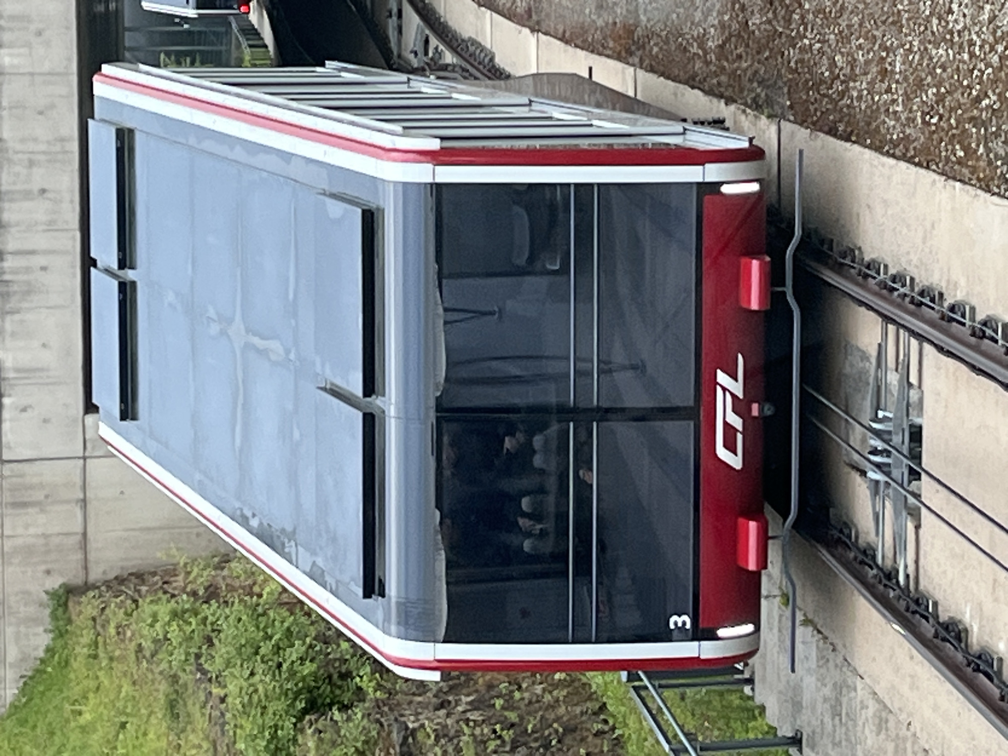 The image shows a red and gray CFL funicular car on a railway track. The funicular car has the number "3" on its front. The environment includes greenery and a concrete wall. The interior of the car appears to have passengers. The scene is likely located in Luxembourg City, Luxembourg, given the presence of the CFL (Chemins de Fer Luxembourgeois) funicular, a public transport system in Luxembourg.