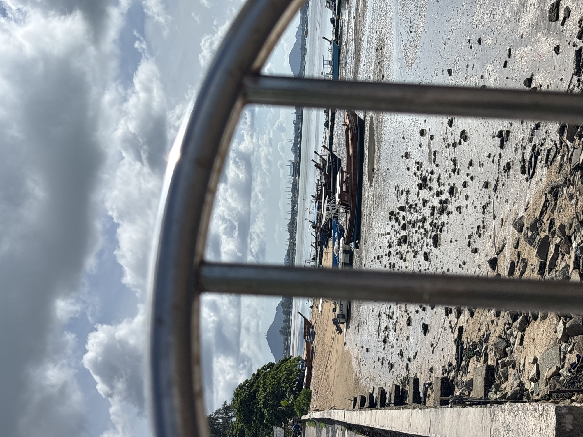 The image depicts a coastal scene at low tide in Rawai, Thailand, viewed through a metal railing. The foreground is dominated by the textured mudflats, littered with rocks, debris, and some scattered seaweed. Several boats are beached or partially submerged in the shallow water and mud. These include traditional longtail boats and some smaller motorized vessels.
Further back, a sandy beach is visible, with more boats resting on it, including a notable wooden boat with intricate details. Trees and vegetation line the edge of the beach, with a road and a low concrete wall separating the beach from the viewpoint. In the background, the sea extends to the horizon, dotted with numerous small boats and structures on the water. A distinctive, tree-covered mountain with antenna-like structures on its peak rises in the distance, characteristic of the Phuket region. The sky is partly cloudy, with a mix of white cumulus and darker grey clouds, suggesting an overcast or partly sunny day. No people are clearly visible in the frame. The overall impression is one of a working harbor or coastline during low tide.