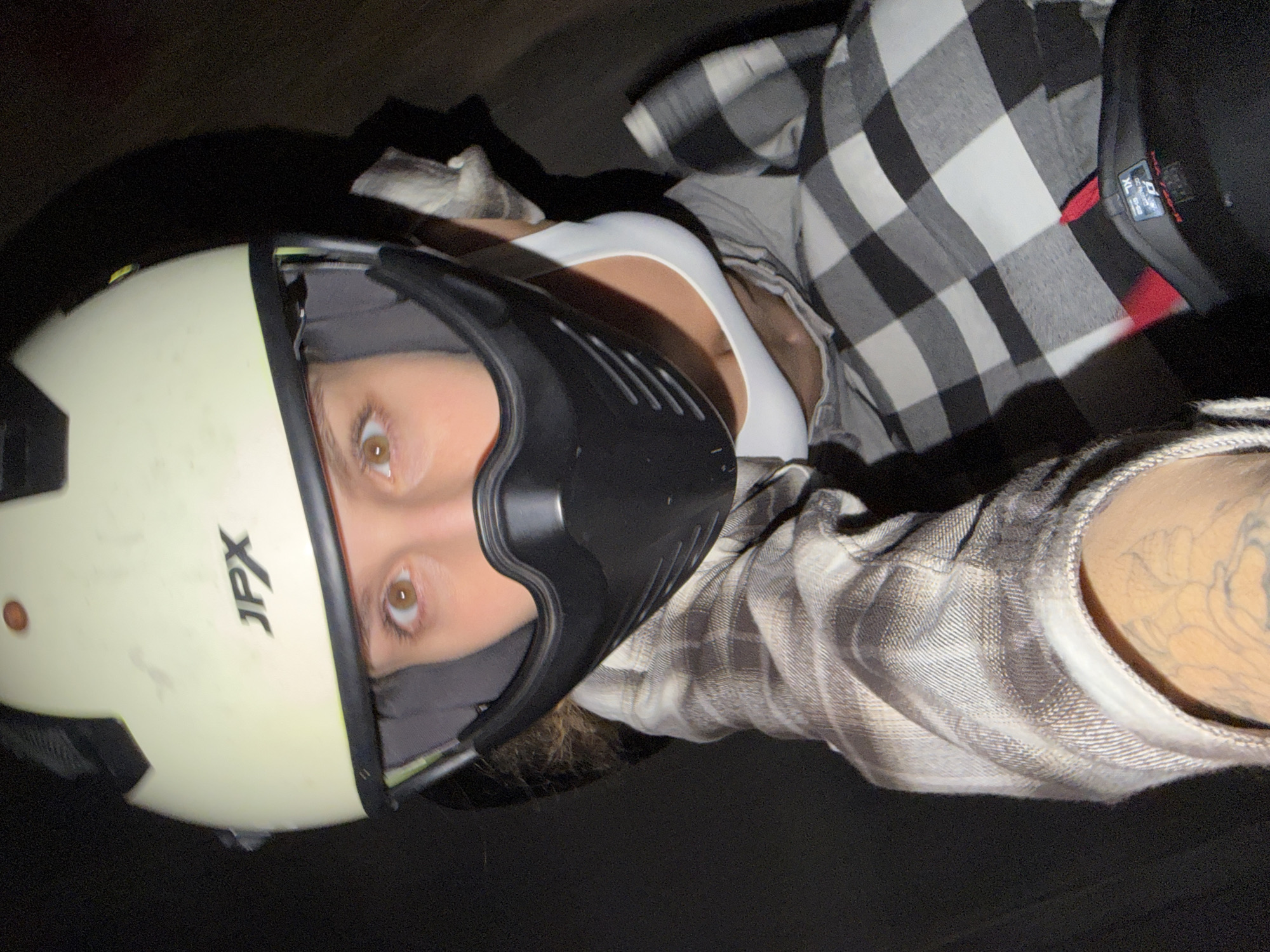 A close-up, low-light portrait captures a person, likely in North Kuta, Indonesia, wearing a light-colored (cream or off-white) helmet with a black integrated face shield or mask. The brand "JPX" is visible on the side of the helmet. The person's wide, brown eyes look directly into the camera, conveying an alert expression. They are dressed in a white tank top or camisole, over which a black and white plaid flannel shirt is worn open. A tattoo is partially visible on the person's left forearm, emerging from the sleeve of the flannel shirt. On the person's right side, beneath the flannel, a black object, possibly a hat or another part of their gear, is visible with an "XL" size label. The background is dark and indistinct, suggesting the photograph was taken at night or in a very low-light setting. The image has a slightly tilted perspective, characteristic of a selfie.