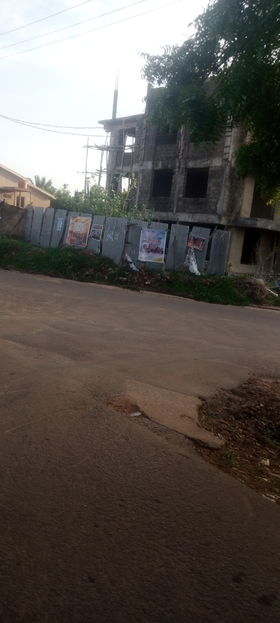The image shows a street scene in Narayi, Nigeria. A two-story building under construction is visible behind a corrugated metal fence. Several posters are attached to the fence. A paved road fills the foreground. A tree is on the right side of the building. Power lines are visible in the sky. Another building is on the left. No people are visible.