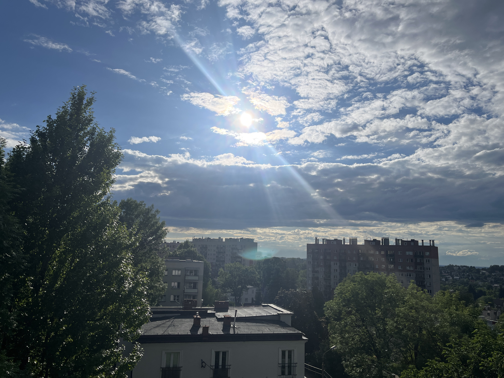 An aerial view of a city skyline under a partly cloudy sky. The sun is visible, shining through gaps in the clouds and casting bright rays across the scene. In the foreground, the tops of several buildings are visible, along with dense green trees. Further in the distance, taller apartment buildings and more trees fill the landscape. The sky is a mix of blue and white, with streaks of sunlight highlighting the clouds.