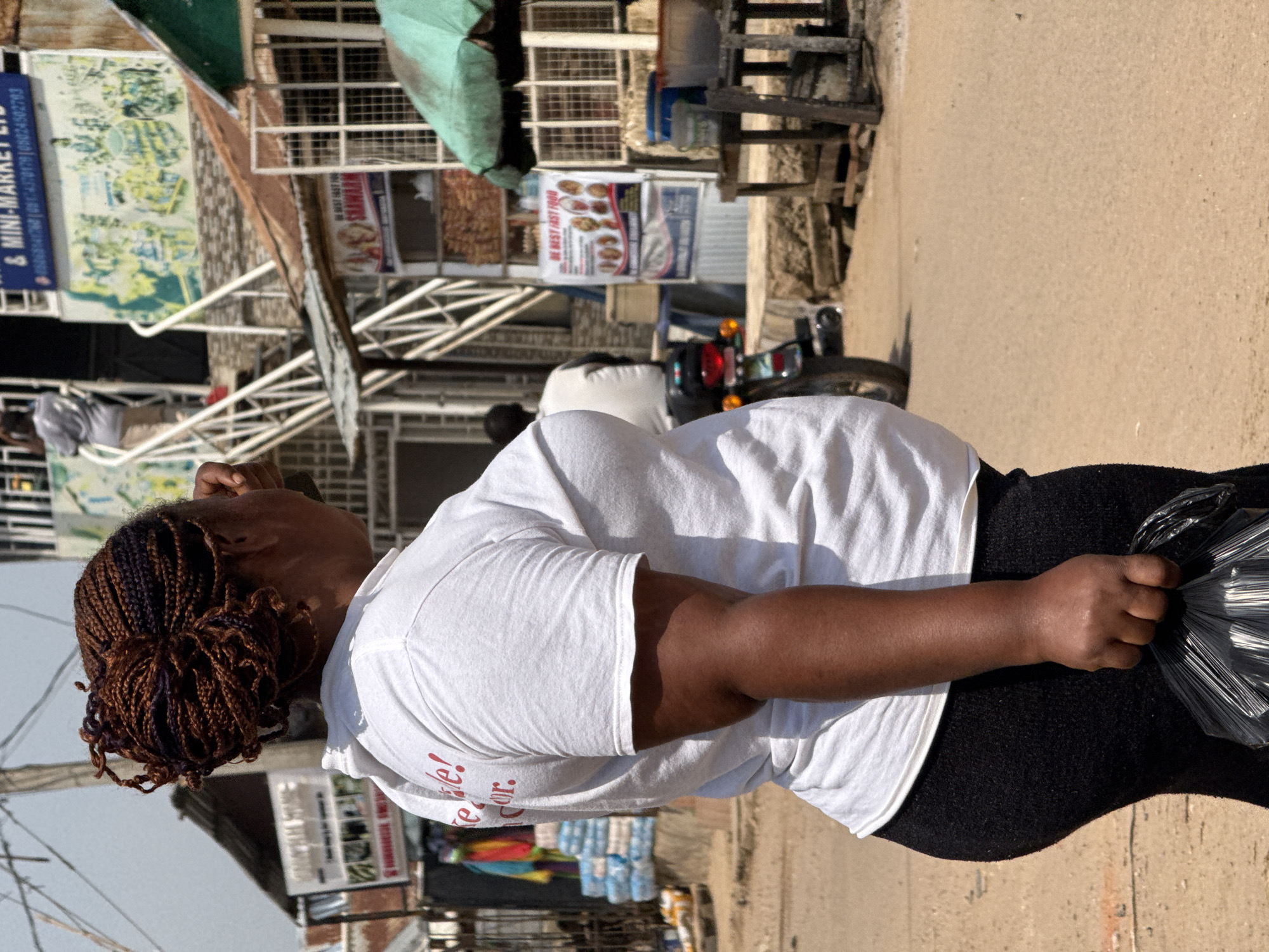In Abuja, Nigeria, a woman with dark skin and braided hair stands in a street market. She wears a white t-shirt with red lettering that reads "Life! For." and black leggings. She is holding a black plastic bag in her right hand. Behind her, a motorcycle is parked. In the background, there are market stalls and buildings, including one with stairs leading up to it. The ground is a dirt road. The overall scene depicts a busy, everyday moment in a Nigerian market.