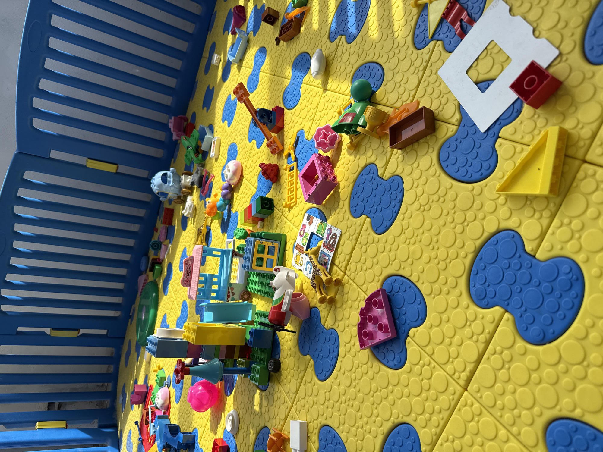 The image depicts a child's play area. A blue and white plastic playpen encloses a section of interlocking yellow and blue foam floor tiles. Numerous plastic toy blocks, including recognizable LEGO bricks, are scattered across the floor, some assembled into rudimentary structures. The lighting suggests an indoor setting with natural light. No people are visible. There is no information about Синьково, Russia in the image.