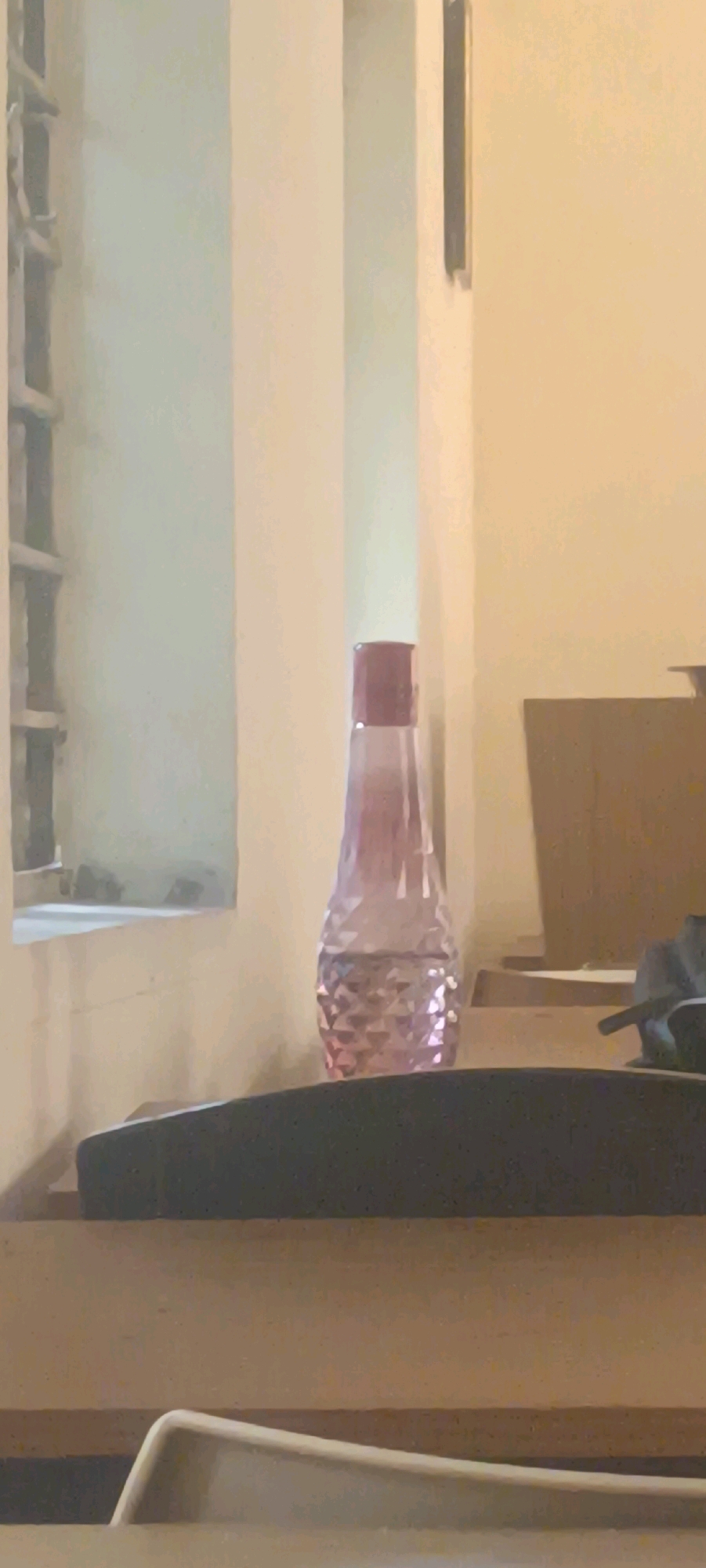 A pink patterned glass bottle with a red cap is centered in the frame, sitting on a dark surface. The bottle contains a clear liquid. Behind the bottle, the background consists of cream-colored walls and a light-colored window with vertical bars on the left. In the foreground, a dark, textured surface, possibly a desk or shelf, is visible. The lighting is soft and diffused, creating a muted atmosphere. The image appears to be taken indoors.