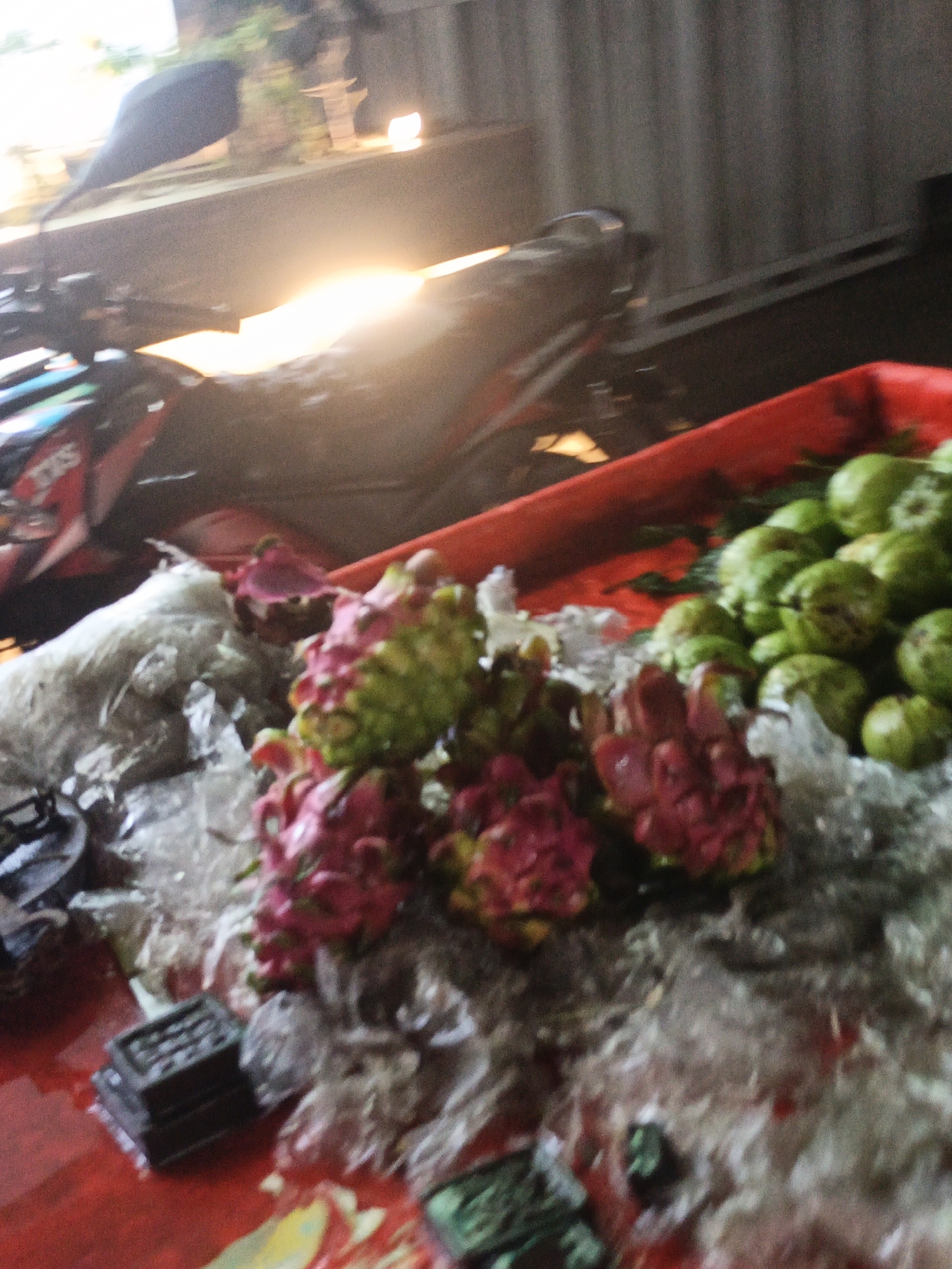 The image depicts a street-side stall in Dhaka, Bangladesh, likely at night or in low light conditions, showcasing a variety of fruits and a motorcycle. In the foreground, a vibrant red tray overflows with fresh produce. Dragon fruits, characterized by their pink and green scaly exteriors, are prominently displayed, some appearing sliced open to reveal their white and pink flesh speckled with seeds. Alongside the dragon fruits are mounds of green guavas. Ice is scattered amongst the fruits, suggesting they are being kept cool for sale. To the left of the fruits, a white, textured material, possibly a bag or cloth, is visible, and beneath it, a small, dark, rectangular object that appears to be a stack of trays or boxes. In the midground, a motorcycle, predominantly black with red accents and the visible "TVS" logo, is parked. Its headlight is illuminated, casting a bright, diffused light across the scene. Behind the motorcycle, a corrugated metal wall forms the backdrop. The overall impression is of a busy marketplace, with the stall and its offerings illuminated by the motorcycle's light and possibly other ambient lighting, creating a dynamic and somewhat hazy atmosphere.
