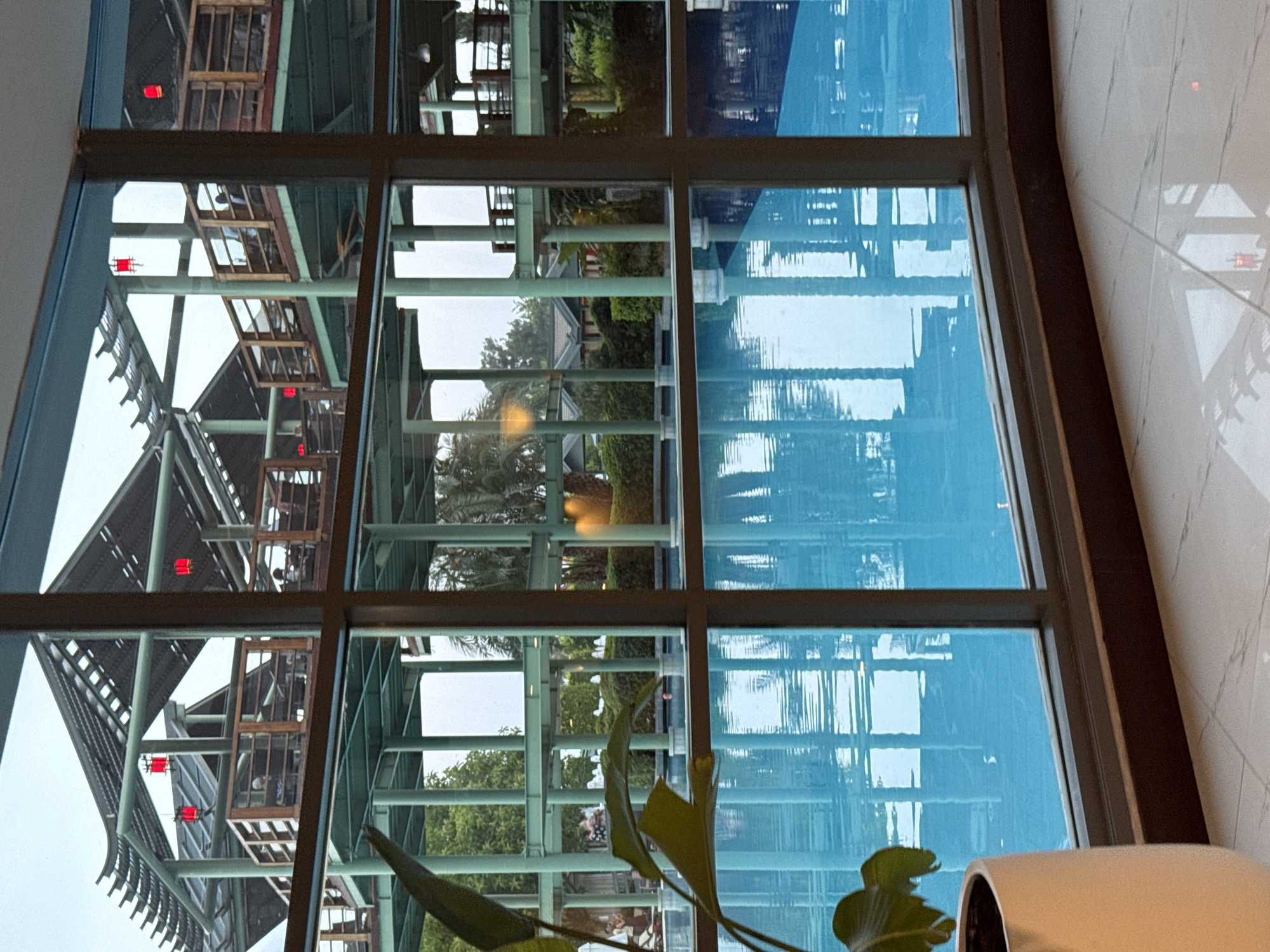 The image shows an indoor swimming pool with bright blue water. The pool is viewed through a large window with a grid of panes. Outside the window, there is a multi-story structure with a pagoda-like roof, supported by green pillars. Red lanterns are visible hanging from the roof. Several people are seated at tables on balconies within this structure. Behind the structure, there are trees and bushes, suggesting an outdoor environment. On the left side of the foreground, a large green leaf from a potted plant is visible, partially obscuring the view. The floor in the foreground is tiled with a marble pattern. The scene appears to be set in a hotel or resort complex. The location is identified as Aksu, Turkey.