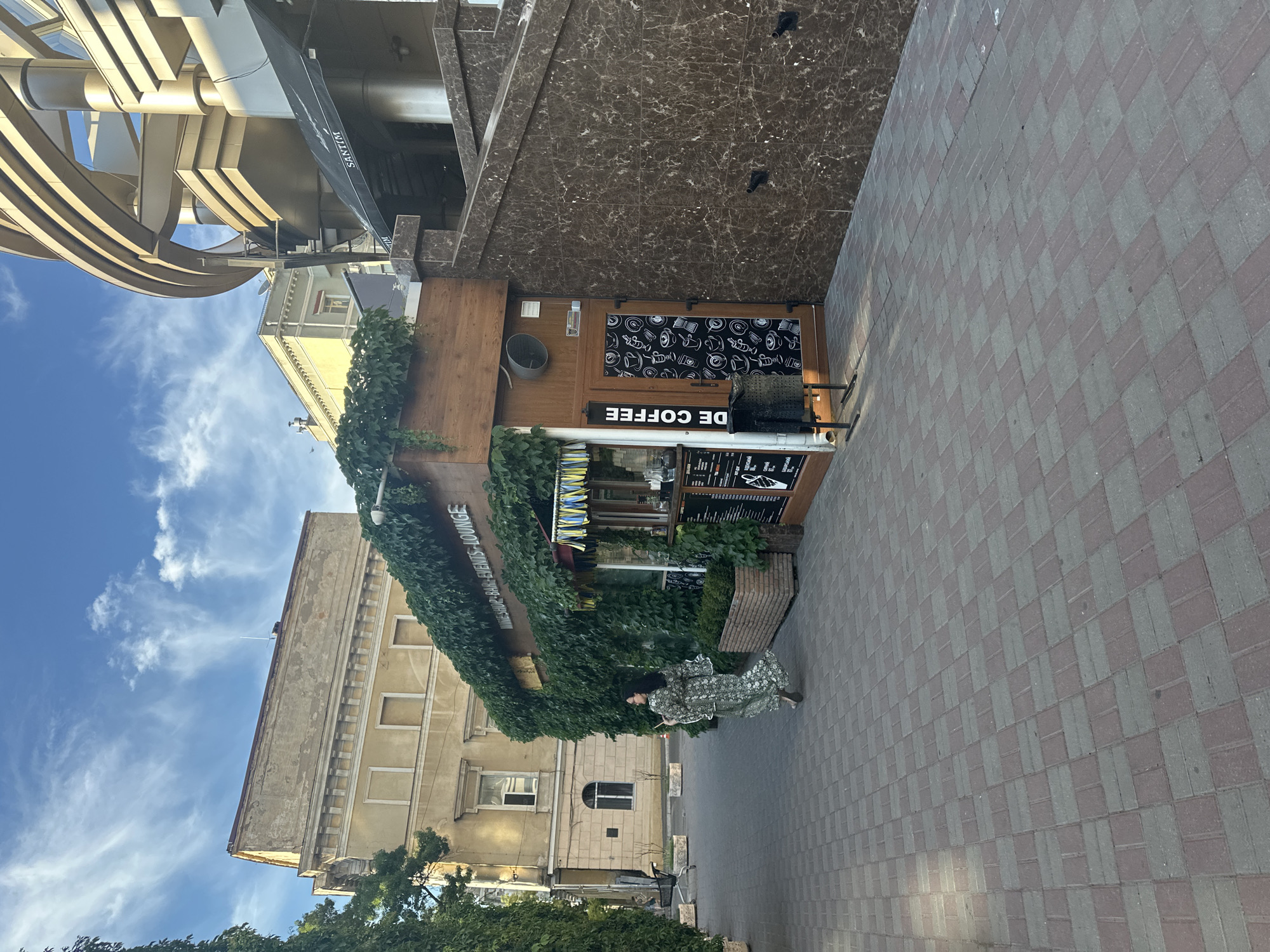 This media file captures an outdoor scene in Odesa, Ukraine. The dominant structure is a small, wooden coffee shop, heavily adorned with lush green ivy, suggesting it's a warm season, likely late spring or summer. A paved walkway, made of alternating red and gray bricks, runs alongside the shop. In the background, a tan-colored building with a classical architectural style, featuring arched windows and decorative cornices, stands tall against a bright blue sky dotted with wispy white clouds. To the left, a modern, metallic structure with curved beams, possibly part of a larger building or an installation, overlooks the scene.
A woman wearing a patterned dress is walking along the paved path, her back partially to the camera. She appears to be holding something small and white in her hand. The overall atmosphere seems peaceful and casual, indicative of a daytime setting.
Visible text on the coffee shop includes "DE COFFEE" written in large, white, reversed letters on a black sign. Another sign above it displays what appears to be a menu or other information, with various items listed. A partially obscured sign on the building behind the coffee shop reads "WINE SHOP - BAR - EVENTS - LOUNGE". There is also text on a black awning of the modern structure on the left, which seems to say "SANTIN". The time of day appears to be late afternoon or early evening, given the angle of the shadows.