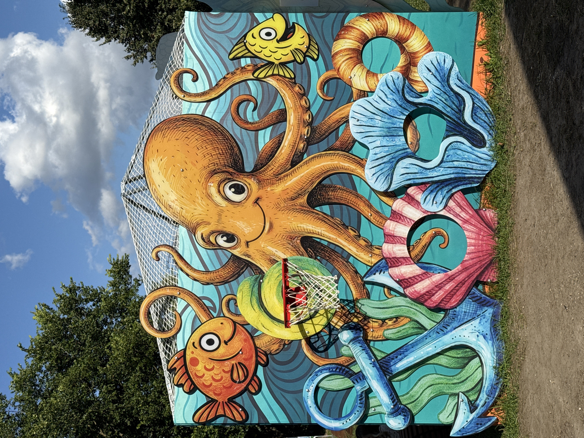 The image shows a colorful, cartoonish mural of an underwater scene that has been turned into a photo booth with cutouts. At the center is a large, smiling orange octopus with its tentacles spread out. To the left, a bright orange fish with large eyes and a wide smile swims towards the octopus. To the right, a smaller yellow fish with a similar expression looks towards the octopus.
In front of the mural, there are several three-dimensional cutouts that guests can pose with. A large blue anchor stands on the left, with green seaweed behind it. In the center, a pink and white seashell cutout has a hole for a face. To the right of the seashell, a blue coral-like cutout also features a face hole. Behind these, a life preserver with orange and yellow stripes also has a cutout.
A basketball hoop with a red rim and white net is positioned within the mural, seemingly in front of a swirling green shape. The background of the mural depicts wavy blue water. Above the mural, a white net structure is visible, possibly part of a canopy or tent. Trees with green foliage are visible on either side of the structure, and a blue sky with white clouds is overhead. The ground in front of the mural consists of dirt and some grass.
This photo booth is located in the city of Khimki, Russia, specifically at the amusement park in the Arkangelskaya Street, building 10, within the "Green Town" residential complex.