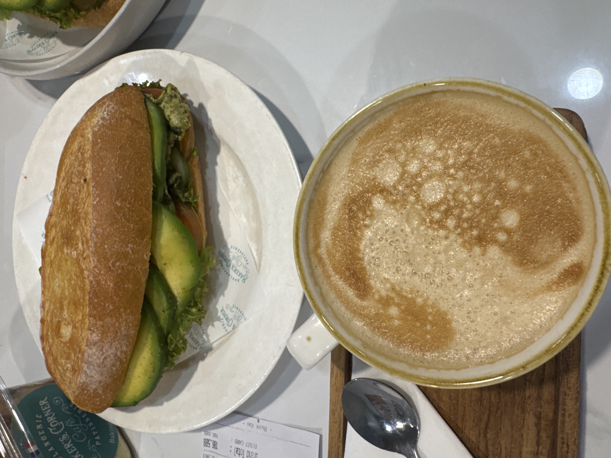 A close-up overhead shot shows a meal at Baker's Corner cafe in Kuta Selatan, Indonesia. In the foreground, a light brown ceramic mug filled with coffee with foamy latte art sits on a white saucer and wooden coaster. A silver spoon rests on a white napkin to the left of the mug. In the mid-ground, a white plate holds a sandwich with a toasted bun, avocado slices, lettuce, and tomato. The plate is adorned with a patterned paper liner that reads "Baker's Corner Cafe Patisserie." A small glass jar with a green label, also featuring the cafe's branding, is visible in the upper left corner, partially obscured by the sandwich. A crumpled receipt is visible in the lower left quadrant. The scene is set against a white table surface, with some reflections visible on the tabletop.