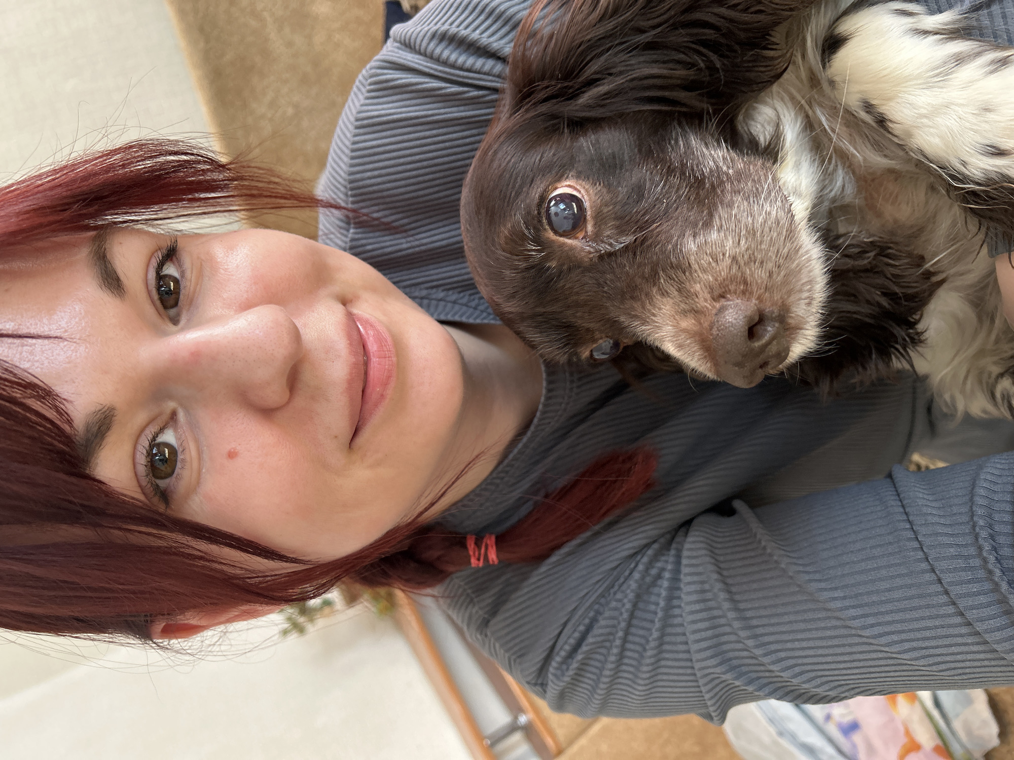 A selfie-style photograph shows a woman with reddish-brown hair and a brown and white dog. The woman is wearing a gray, long-sleeved shirt. Her hair is partially pulled back with a red hair tie. She is looking at the camera and smiling slightly. The dog is positioned to the right of the woman, also facing the camera. The dog's fur is predominantly brown, with white patches around its face and paws. The location is likely indoors, with a glimpse of a wooden door frame and furniture in the background. The image does not provide enough detail to establish a specific location within the city of Фоки, Russia.