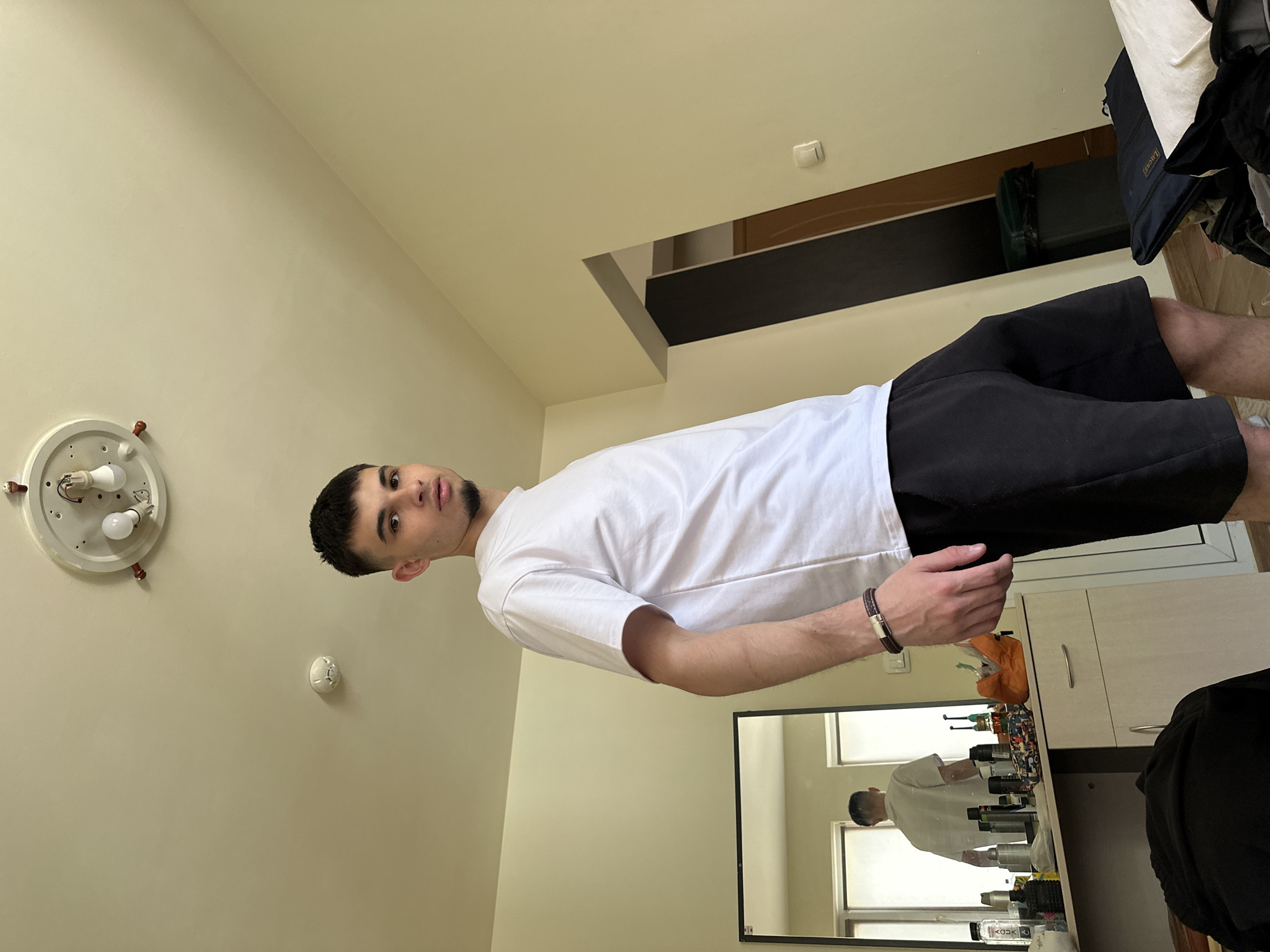 The image depicts a young man standing in a room. He has dark hair and a light beard. He is wearing a white t-shirt and black shorts. He is standing next to a dresser with a mirror. The dresser has various items on it, including toiletries and grooming products. The room is painted in neutral tones. There is a light fixture on the ceiling. The location and the city of Sofia, Bulgaria cannot be determined from the image.