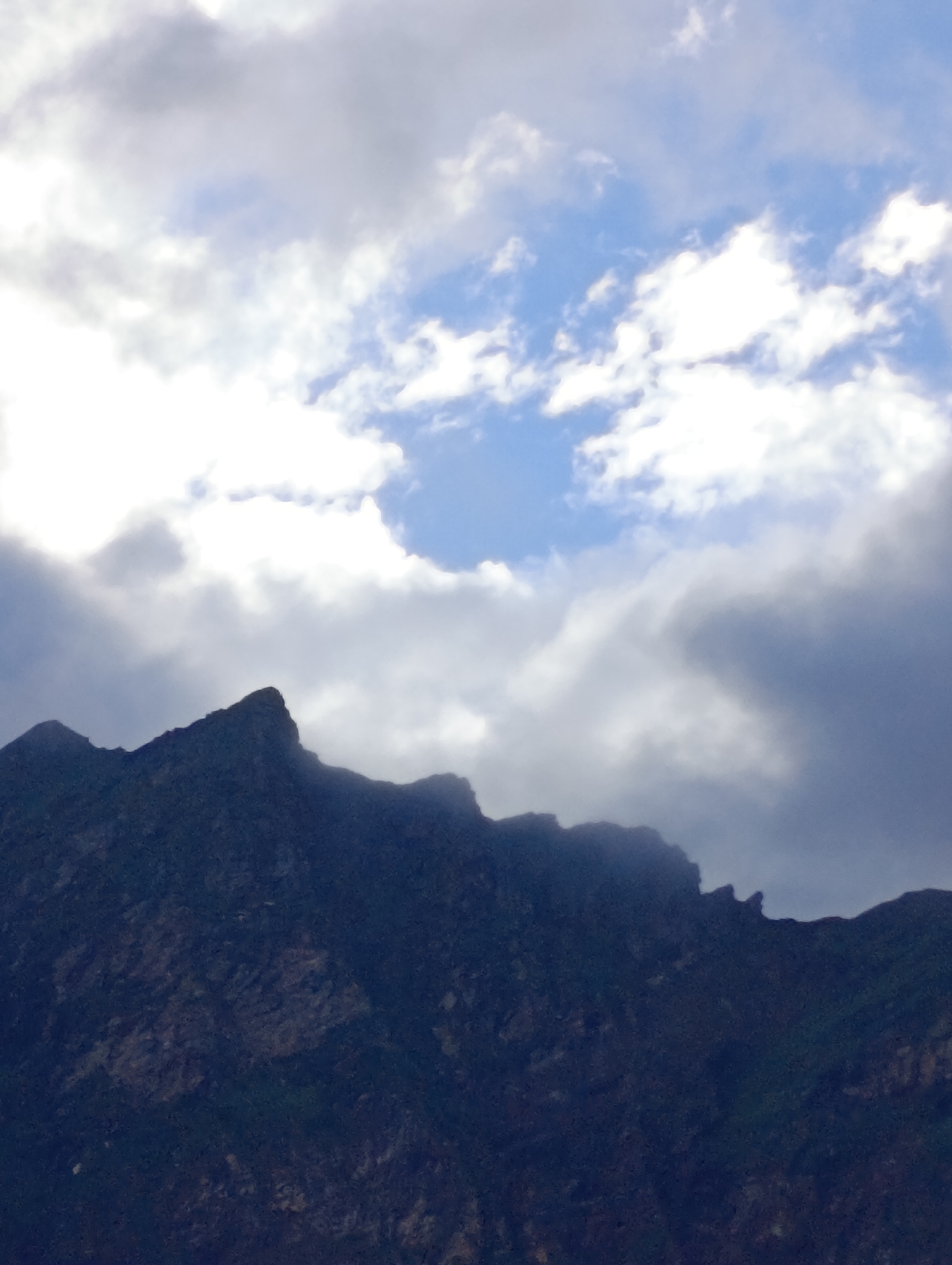 This is a high-angle, medium shot of the jagged peaks of a mountain range against a cloudy sky in Marhi, India. The mountain is dark and silhouetted, with a rough texture visible on its surface. The sky is a mix of bright white and grey clouds, with patches of blue sky showing through. The lighting is diffused, suggesting an overcast or partly cloudy day. There are no people or man-made objects visible in the image. The environment appears to be a natural, mountainous landscape.