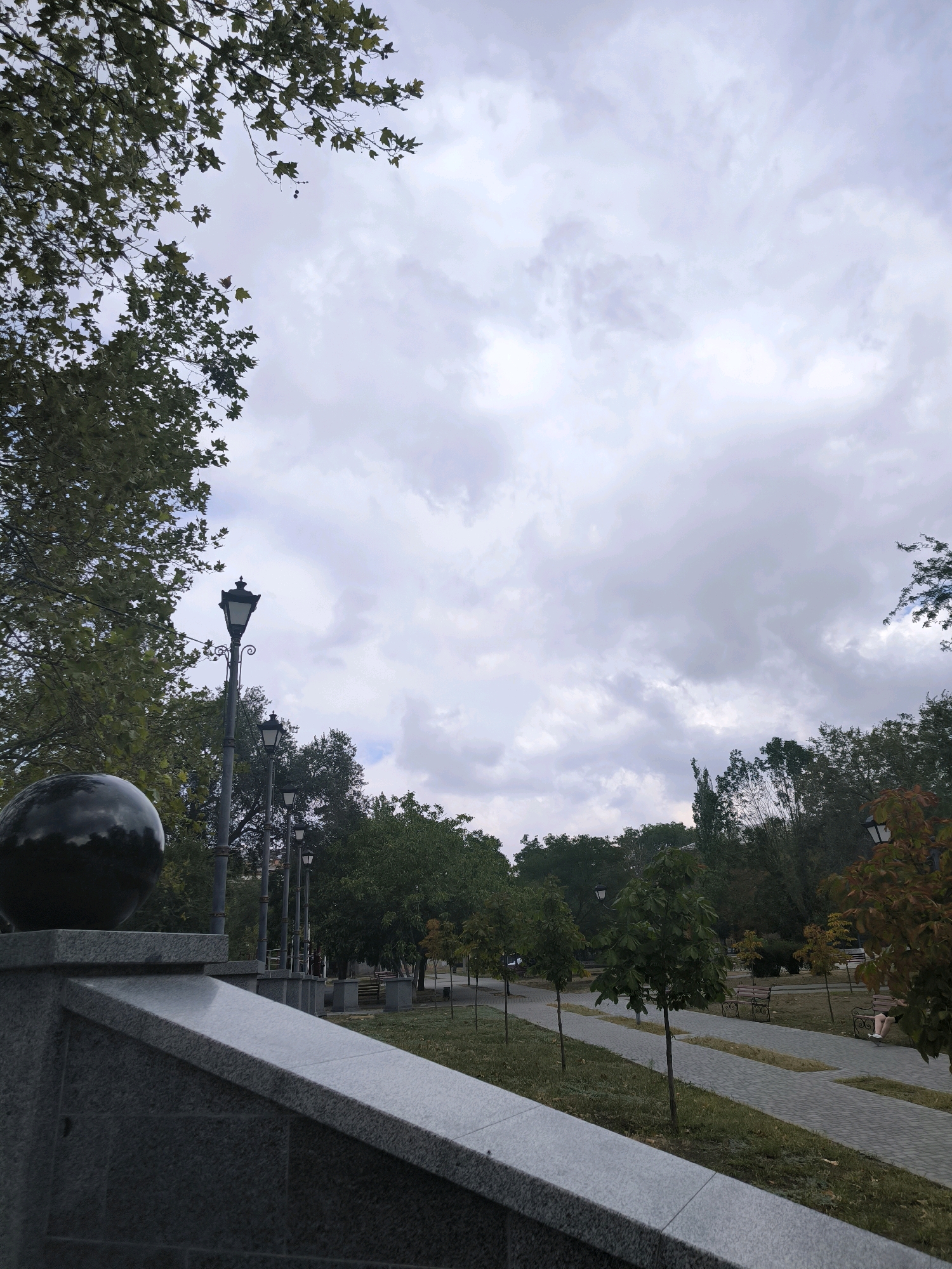 The image depicts a view of a park pathway in Mykolaiv, Ukraine. A granite embankment with a dark spherical ornament is in the foreground on the left. Beyond the embankment, a paved pathway lined with young trees and park benches runs diagonally through the scene towards the right. Several ornate lampposts are visible along the pathway, with taller ones on the left and shorter ones further down. Lush green trees with some autumn foliage are present on both sides of the pathway, and a person is seated on a bench on the right. The sky is overcast with heavy clouds.