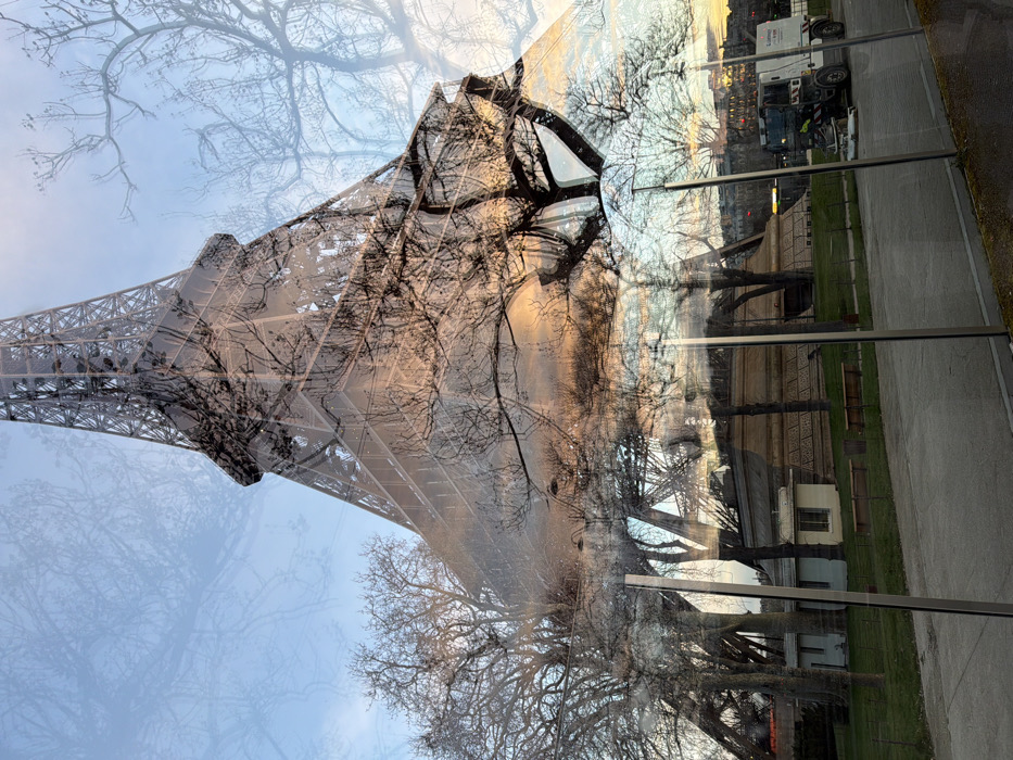 The image displays a complex composite scene, primarily a reflection on a glass surface, centered on the Eiffel Tower in Paris, France. The upper two-thirds of the image show the distinctive steel lattice structure of the Eiffel Tower, partially obscured and overlaid by the stark, intricate silhouettes of numerous bare deciduous tree branches against a sky exhibiting a gradient from light blue at the top to warm orange and pink hues towards the horizon, indicating either sunrise or sunset.
The lower portion of the image, visible through and reflecting on the foreground glass, depicts a park-like environment. A paved path or road horizontally traverses this section. To the left of the path, a small, light-colored, single-story building with a darker roof is situated alongside a larger, multi-tiered stone structure. Several bare trees are rooted in the green grassy area, which also contains two unoccupied wooden benches. To the right of the path, a white and grey street sweeper vehicle with green accents and a "Groupe C" logo is positioned on the paved surface, appearing stationary. Vertical, silver-colored metallic frames of the foreground glass barrier are visible, contributing to the layered visual effect. No individuals are present in the scene.
