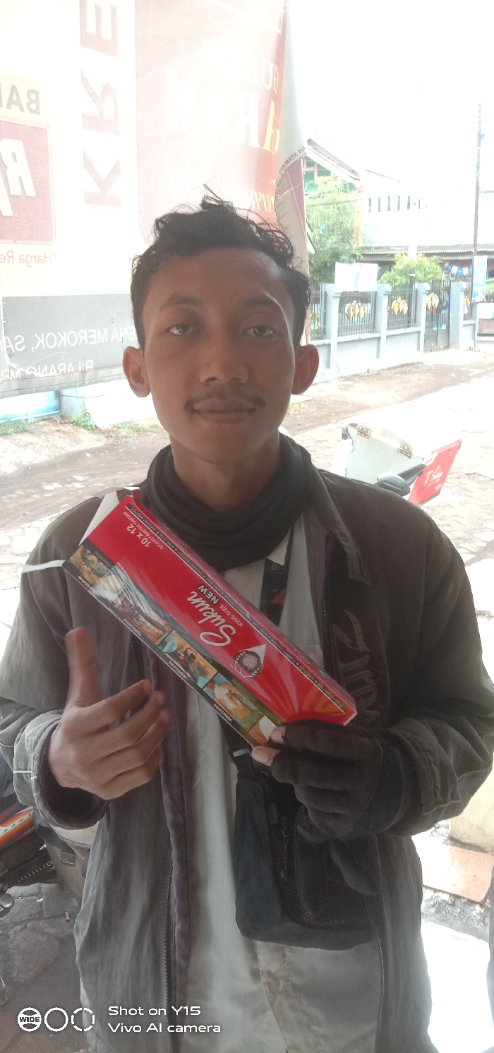 The image depicts a young adult male, likely Indonesian, standing outdoors in the city of Sebaung, Indonesia. He has short dark hair and a thin mustache, looking directly at the camera with a neutral expression. He is wearing a dark brown or olive-green zipped jacket over a lighter garment and a black neck gaiter. His right hand is covered by a black glove.
The male is holding a large, red, rectangular carton, likely a retail display box of cigarettes, in both hands, angled upwards to his left. The visible side of the carton is labeled "Subur King Size NEW 10 x 12" and features graphic health warnings and product imagery. His left thumb is partially visible, giving a "thumbs up" gesture.
In the background, a large, faded white banner with red text, partially mirrored, is visible on the left, displaying Indonesian words such as "BAHAYA MEROKOK" (Danger of Smoking) and "KRETEK." To the right of the subject, the rear section of a red and white scooter or motorcycle is partially visible. Further back, a gray concrete wall adorned with multiple yellow, leaf-shaped ornaments can be seen. Beyond the wall, a residential building with a red roof and green accents, partially obscured by foliage, is present. The ground appears to be a mix of dirt and unpaved surface. The lighting suggests an overcast daylight condition.