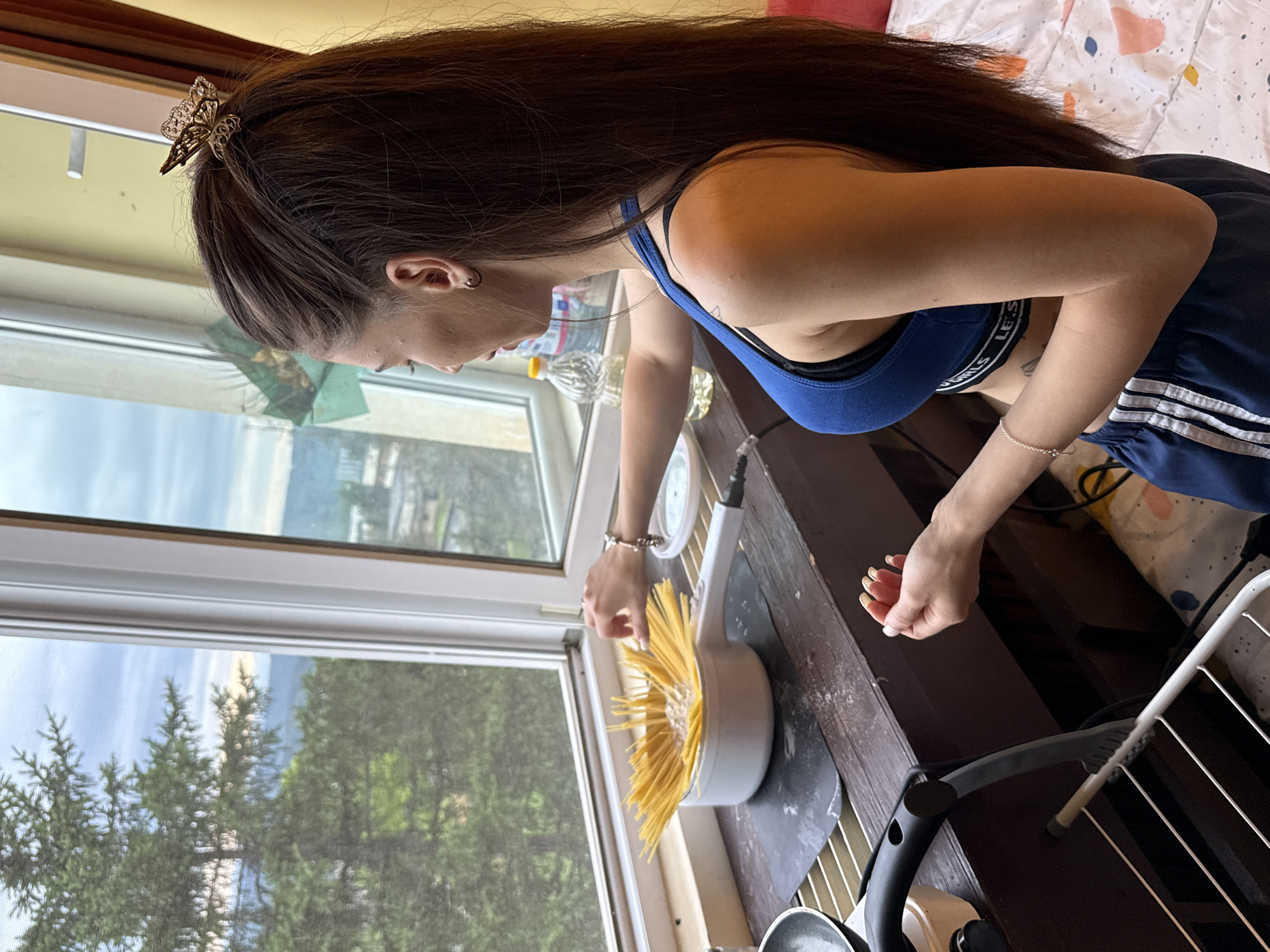A woman with long dark hair, wearing a blue sports bra and dark blue athletic shorts, stands by a window preparing spaghetti. She is in an indoor setting, possibly a kitchen or dining area, with a view of trees and a distant cityscape through the window. A pot of uncooked spaghetti is in the foreground, standing upright in a white pan, with water and possibly some other ingredients in the pan. A bottle of cooking oil and other kitchen items are visible on the windowsill. The woman is holding a piece of spaghetti in her right hand, seemingly about to add it to the pot. Her left hand is also near the pot. A clothes rack is visible in the lower left portion of the image.