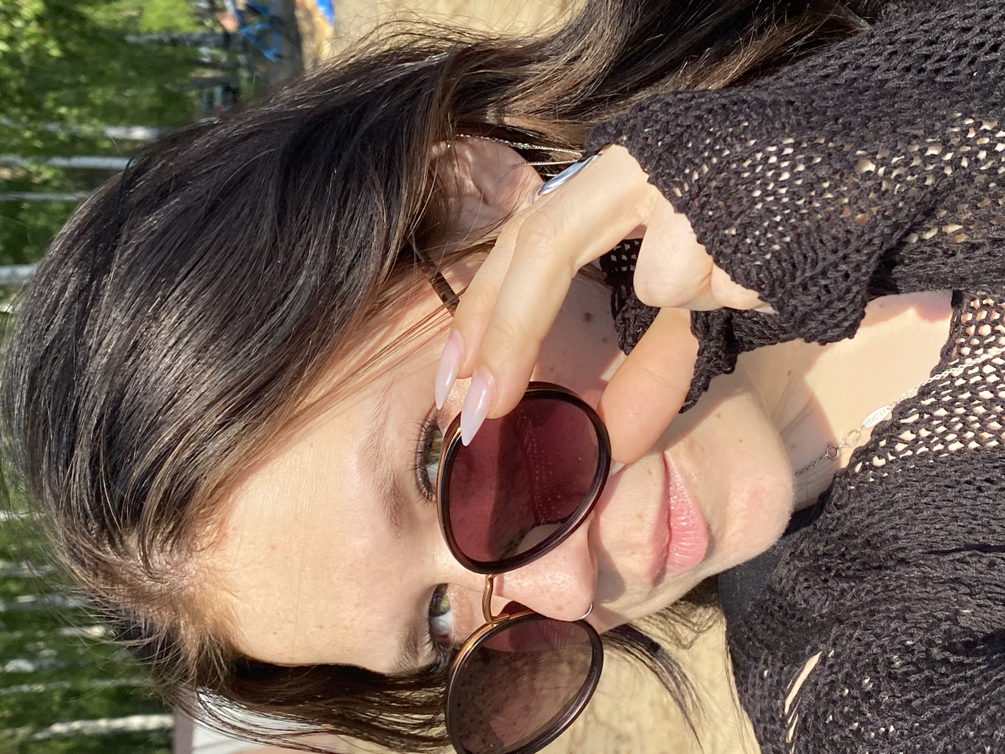 A woman poses outdoors, holding dark-rimmed sunglasses in front of her face. She has long, dark hair, fair skin, and long manicured nails. She is wearing a black, knitted sweater. A thin silver necklace is visible around her neck. The background suggests an outdoor setting, potentially in a sunny area with green foliage in the distance. The location cannot be verified as Chaikovsky, Russia based on the visual information provided.