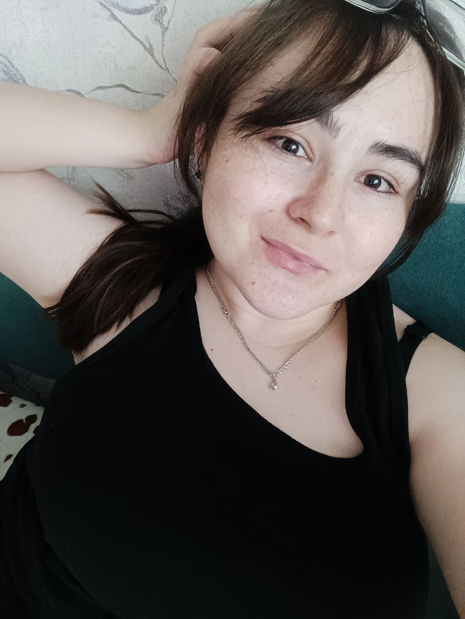 A woman is lying down, taking a selfie. She has dark hair with bangs, and a light complexion with freckles. She is wearing a black tank top and a silver necklace with a pendant. Part of a patterned pillow or cushion is visible to the left, and part of a blue sofa or cushion is visible to the right. The location is unknown.