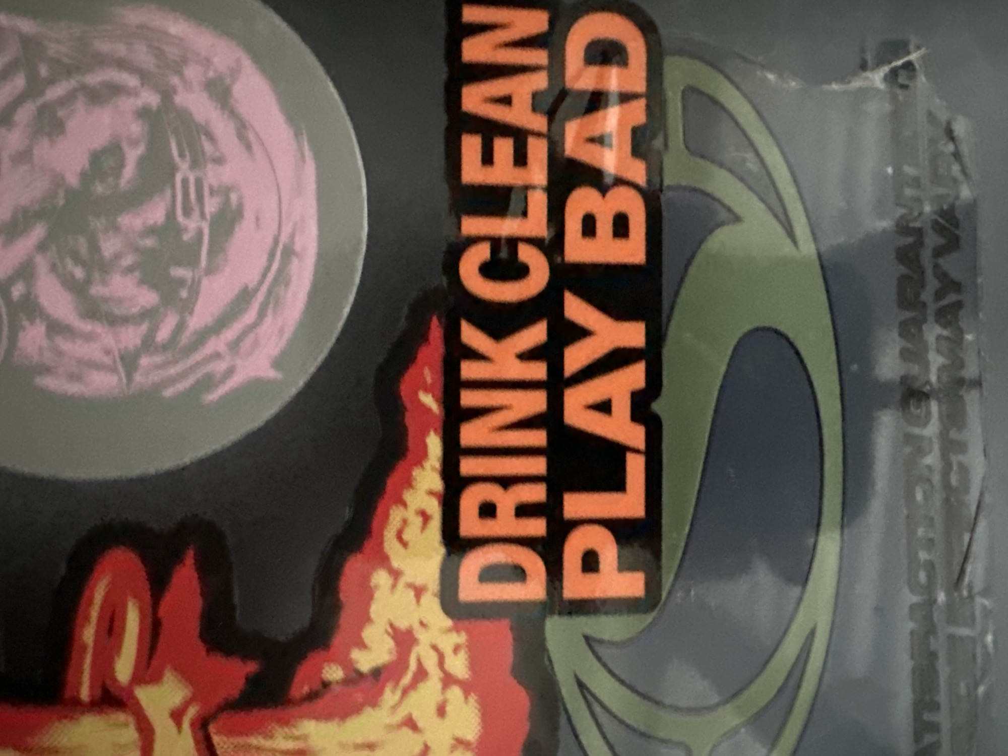 The image displays a close-up view of a graphic design surface, located in the city of Wealden, United Kingdom. The central element is a rectangular orange text box with a black outline, presenting the words "DRINK CLEAN PLAY BAD" in uppercase letters. To the upper left of this text, a red and yellow graphic resembling flames is visible against a dark background. To the upper right, a circular graphic features swirling patterns in pink and grey. Below the central text, a partially visible green-outlined abstract shape or logo is present on a dark grey surface. Towards the bottom, faint, smaller text fragments, including "SATISFACTION GUARANTEE" and "MAY VARY", are discernable. The depicted surface shows minor signs of wear, including slight creases and superficial scratches. No individuals or active interactions are depicted.