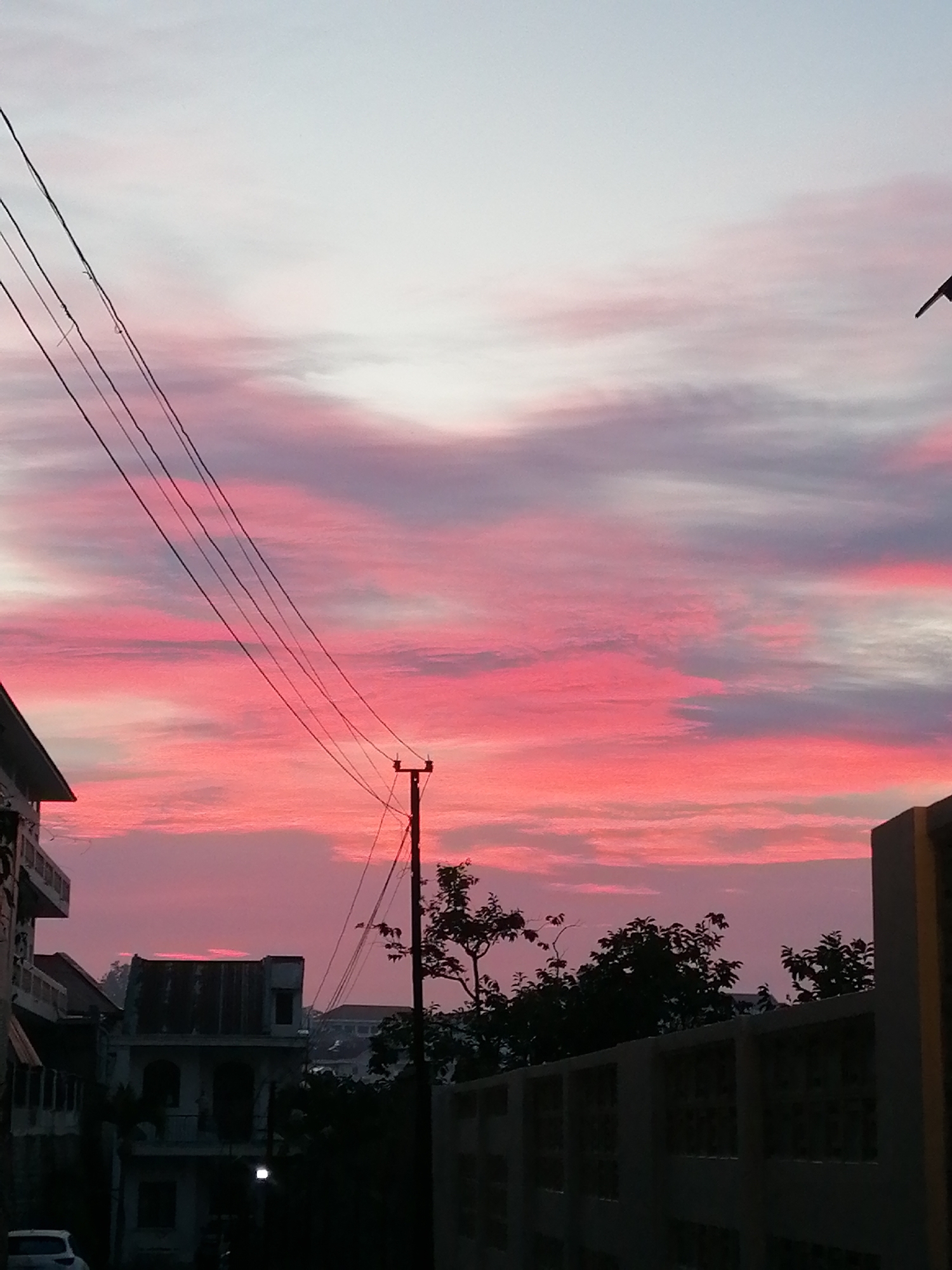 The image shows a cityscape at dusk in Da Lat, Vietnam. The sky is filled with pink and gray clouds, indicating either sunrise or sunset. Power lines stretch across the upper left portion of the image, connecting to a utility pole. In the foreground, the outlines of buildings are visible, including one with a corrugated metal roof. Foliage from trees obscures some of the buildings. Part of a wall is also visible on the right side of the image. A single light source illuminates one of the buildings. A car is visible at the bottom left corner.