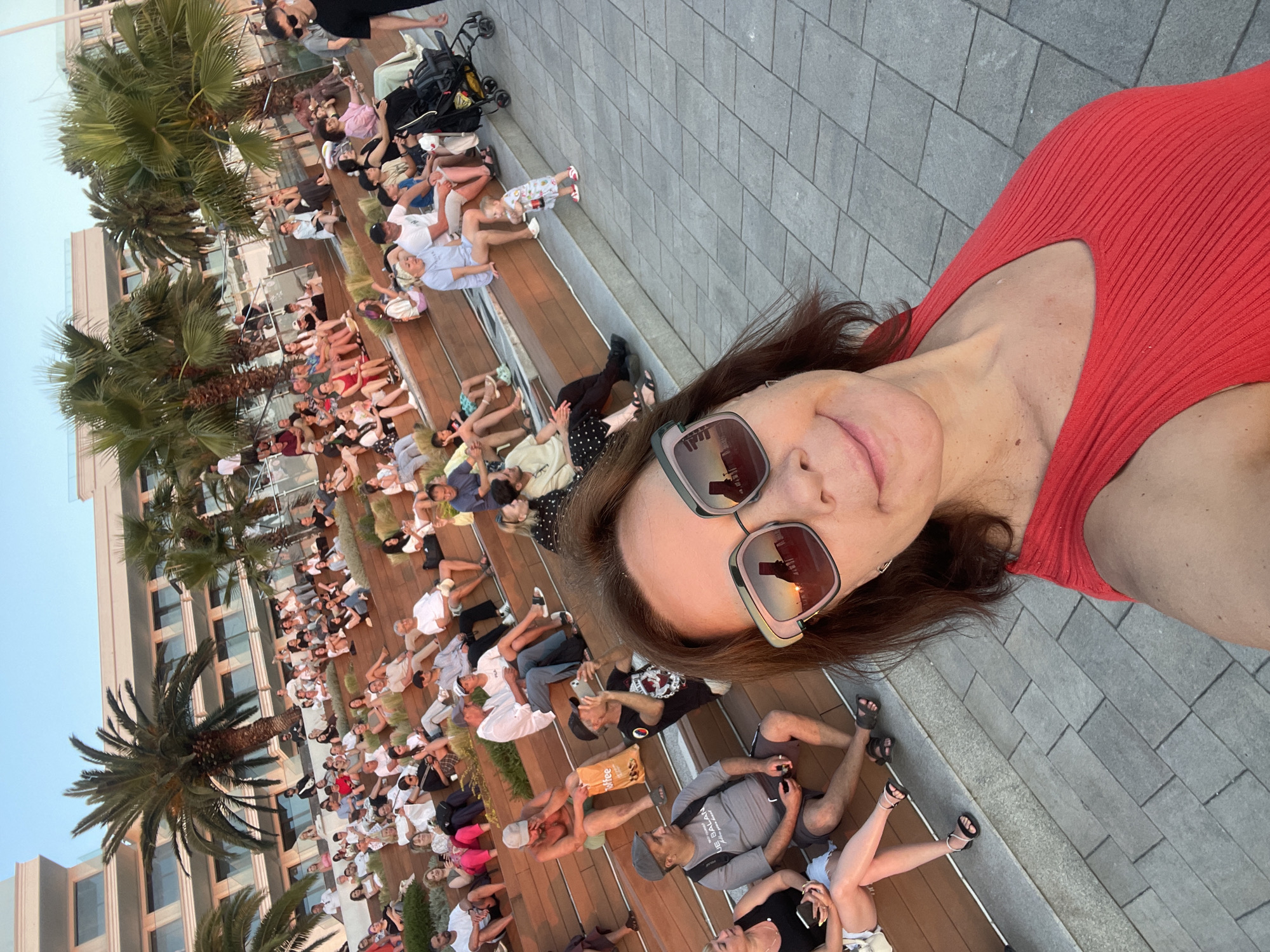 This is a wide-angle, selfie-style photograph taken outdoors in Sochi, Russia. The main subject is a woman in a red top and sunglasses, smiling and looking at the camera. She is in the foreground, occupying the right side of the frame.
Behind her, a large group of people are seated on tiered wooden benches, facing away from the camera. They appear to be watching an event or performance, though the specifics are unclear. Many are casually dressed, and some are holding phones, suggesting they might be recording or taking pictures. The crowd extends down the benches, creating a sense of a gathered audience.
Palm trees are visible on the left side of the image, with their fronds reaching towards the top of the frame, indicating a warm climate. Behind the seated crowd, a modern building with glass windows can be seen, suggesting an urban or resort setting. The ground where the people are sitting is a mix of wooden planks and grey stone paving.
The lighting suggests it is late afternoon or early evening, with a warm glow from the sun, potentially a sunset given the reflections in the woman's sunglasses. The overall mood seems relaxed and communal, with people enjoying an outdoor gathering. There is no visible text in the image.