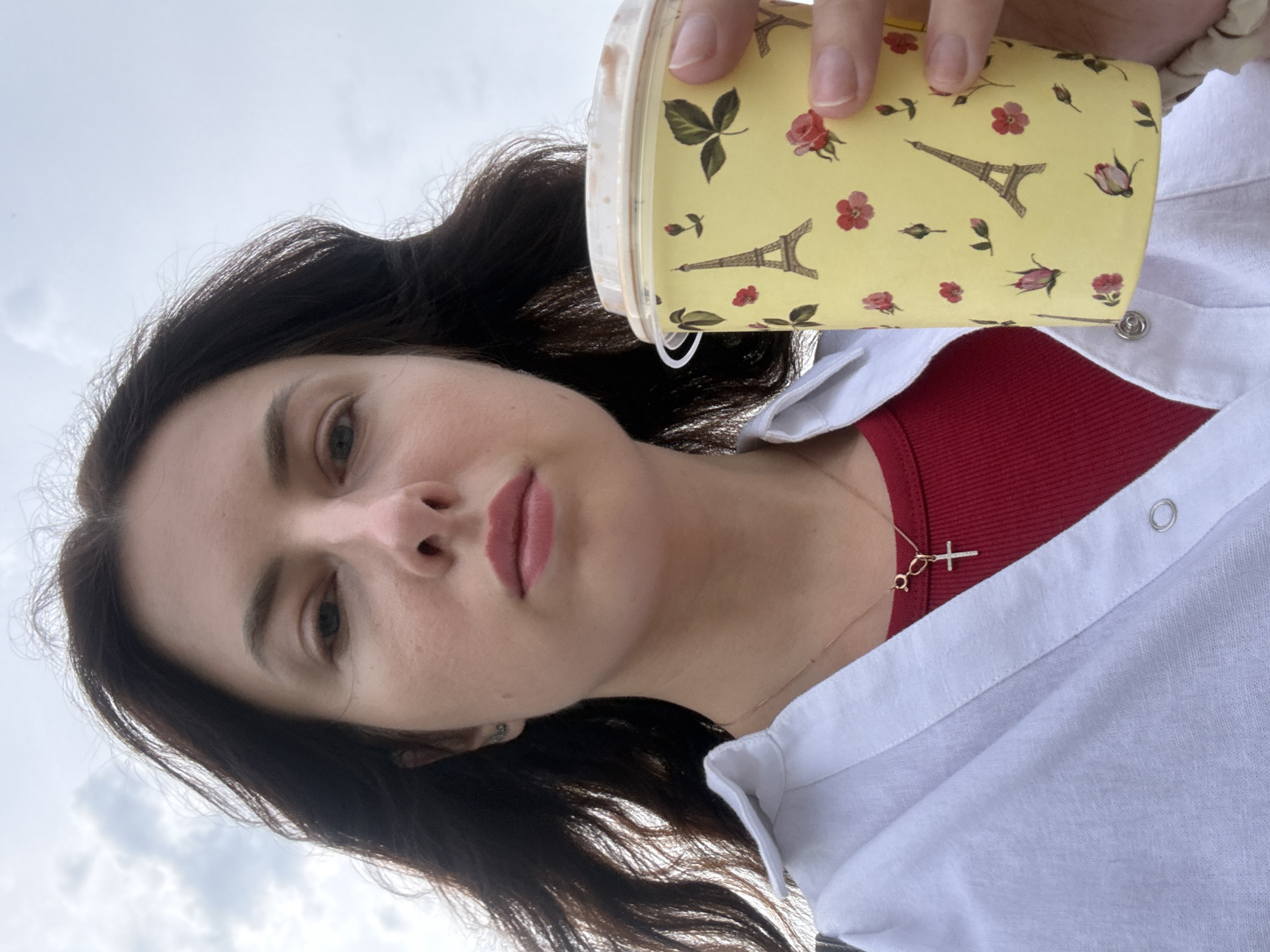 A medium shot captures a woman holding a yellow disposable cup decorated with Eiffel Towers and roses. She is wearing a white unbuttoned shirt over a red top and a delicate gold necklace with a cross pendant. The woman's dark hair is windswept, framing her face as she looks towards the camera. The background consists of a cloudy sky, suggesting an outdoor setting, potentially in Moscow, Russia, given the user's prompt. The lighting is diffused, indicative of an overcast day.