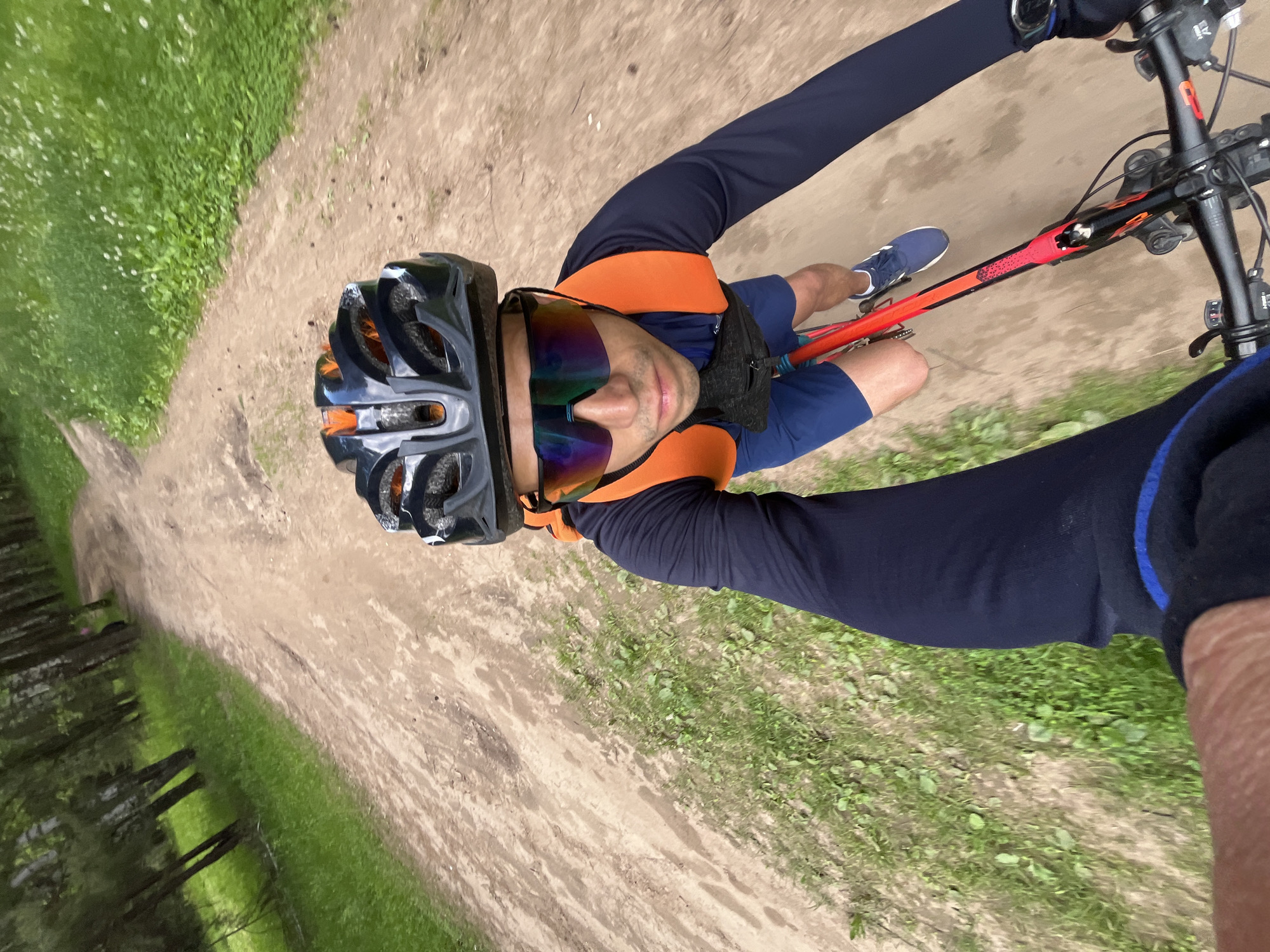 A first-person perspective shot shows a male cyclist riding on a dirt path through a forest. The cyclist is wearing a black helmet, reflective sunglasses, a navy long-sleeved shirt, and blue shorts. He has an orange hydration pack on his back and a small black bag around his waist. The bicycle is red and black. The path is surrounded by green grass and trees. The image is taken in Chaikovsky, Russia.