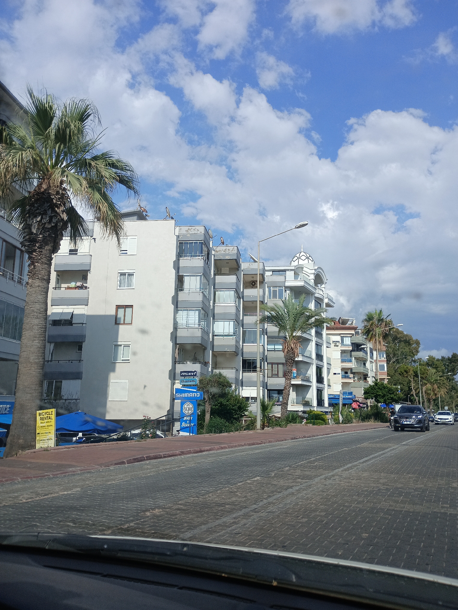 This media file captures a view of a street scene in Alanya, Turkey. The foreground is dominated by the textured cobblestone of the road, with the hood of a vehicle visible at the very bottom. A prominent palm tree stands to the left, its fronds reaching towards a bright blue sky dotted with fluffy white clouds.
Behind the palm tree, a multi-story building with a light-colored facade and grey balconies is visible. Further down the street, other apartment buildings line the road, some with distinctive architectural features like a domed roof. Several cars are driving on the road, including a dark-colored SUV and a white sedan.
Signs for businesses are visible along the buildings. These include "BICYCLE RENTAL," "KRALK BILET," "FIX BIKE," and "SHIMANO." Other signs, partially obscured, mention "MARKET." The presence of a blue umbrella and what appears to be a rental kiosk suggests commercial activity related to tourism or recreation.
The lighting and the presence of shadows indicate it is daytime, likely late morning or afternoon. The weather appears pleasant with a mix of sun and clouds. The overall scene suggests a bustling urban environment, possibly a coastal city, with shops and residential buildings along a main thoroughfare. There are no people prominently featured in the image.