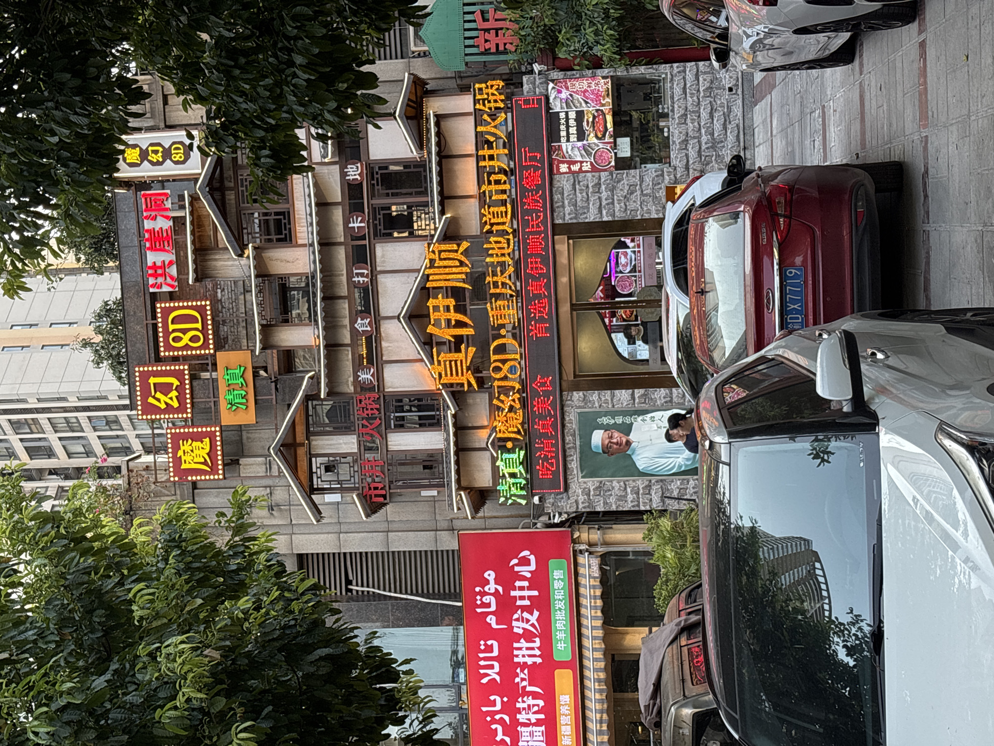 This image, taken in Longxi Subdistrict, China, depicts a bustling street scene with a focus on a multi-story building adorned with numerous illuminated signs, predominantly in Chinese characters. The building appears to be a restaurant or a commercial establishment, indicated by the signage "真伊顺" (Zhen Yi Shun), "吃重庆火锅" (Eat Chongqing Hotpot), and "新疆营养馕" (Xinjiang Nutritious Naan). There are also signs advertising "8D" experiences, suggesting some form of entertainment or unique offering.
In the foreground, several cars are parked along the sidewalk, with a prominent red car featuring a license plate that reads "D-X7719". A large red banner on the building displays text in both Chinese and what appears to be Arabic script, with Chinese characters translating to "Xinjiang Specialty Wholesale Center" and mentioning "beef and lamb wholesale and retail". A framed picture on the building shows a man in traditional attire, possibly a community leader or a representation of the restaurant's ethnic focus.
Lush green trees partially obscure the building, suggesting a temperate climate and possibly a daytime setting, although the bright neon signs create a vibrant, almost nighttime atmosphere. The architecture of the building is varied, with a mix of stone facades and wooden decorative elements, giving it a somewhat traditional yet commercial appearance. The overall impression is one of a lively urban environment with a strong emphasis on food and commerce, particularly highlighting Xinjiang cuisine and products.