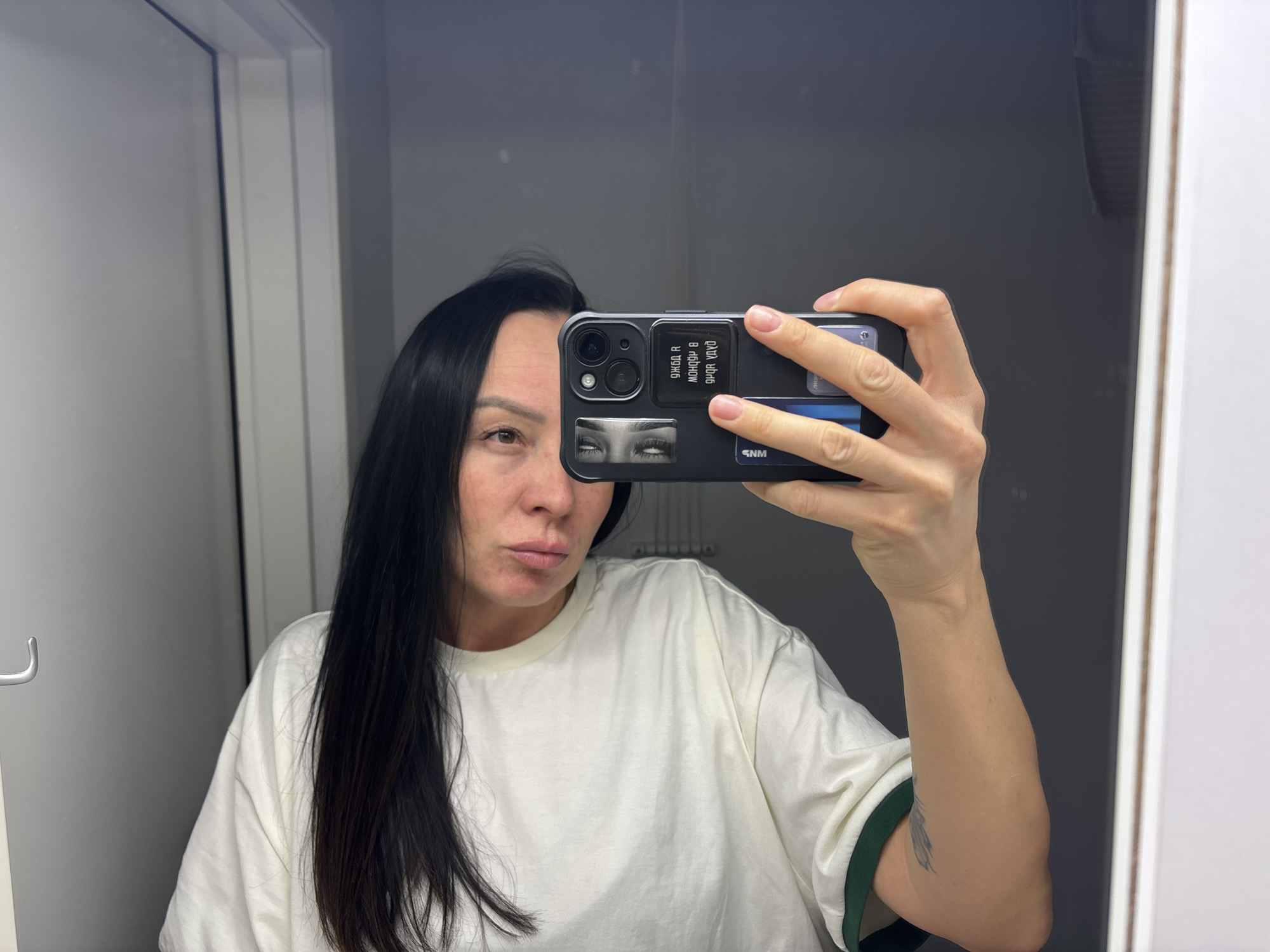 A woman is taking a selfie in what appears to be a bathroom. She is holding a smartphone in her right hand, and the phone partially obscures her face. The phone case has a set of stylized eyes and Russian text. She has long, dark hair and is wearing a white t-shirt with green trim. The bathroom is simple, with a white door and a gray wall behind her. The location is specified to be in the city of Chaikovsky, Russia.