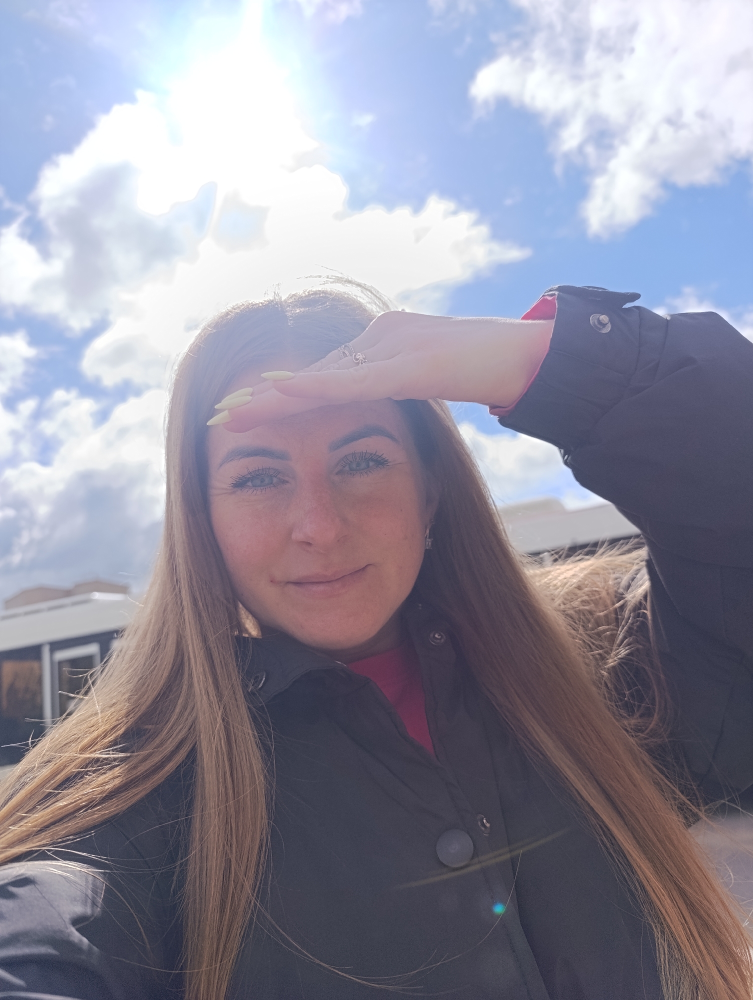 A woman is posing for a selfie outdoors. She is shielding her eyes from the sun with her hand. She has long brown hair and is wearing a dark jacket with a pink shirt underneath. The background shows a bright blue sky with scattered white clouds, and the sun is positioned behind the clouds. A building is partially visible in the background on the left side. The location is an urban environment in the city of Unknown, Unknown.