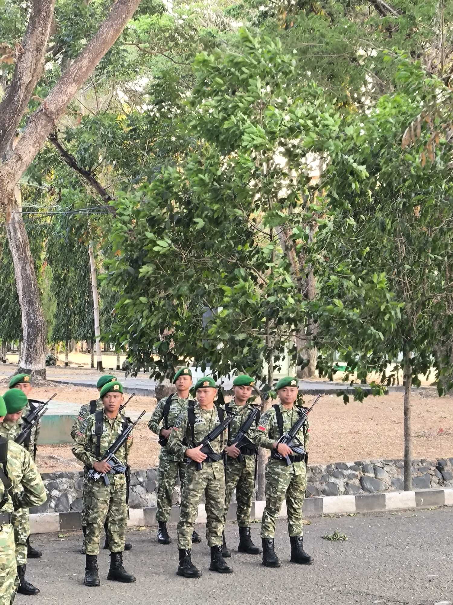 In Unknown, Indonesia, a group of soldiers stands in formation on a paved surface, possibly a road or training ground, with a backdrop of lush green trees and vegetation. The soldiers, all male, are dressed in camouflage uniforms and green berets. They are equipped with rifles, held at the ready or slung over their shoulders. The soldiers are positioned in two rows, with the front row slightly closer to the camera. The lighting suggests it is daytime, possibly late afternoon, given the soft shadows. The environment appears to be an outdoor military installation or a park with trees.
