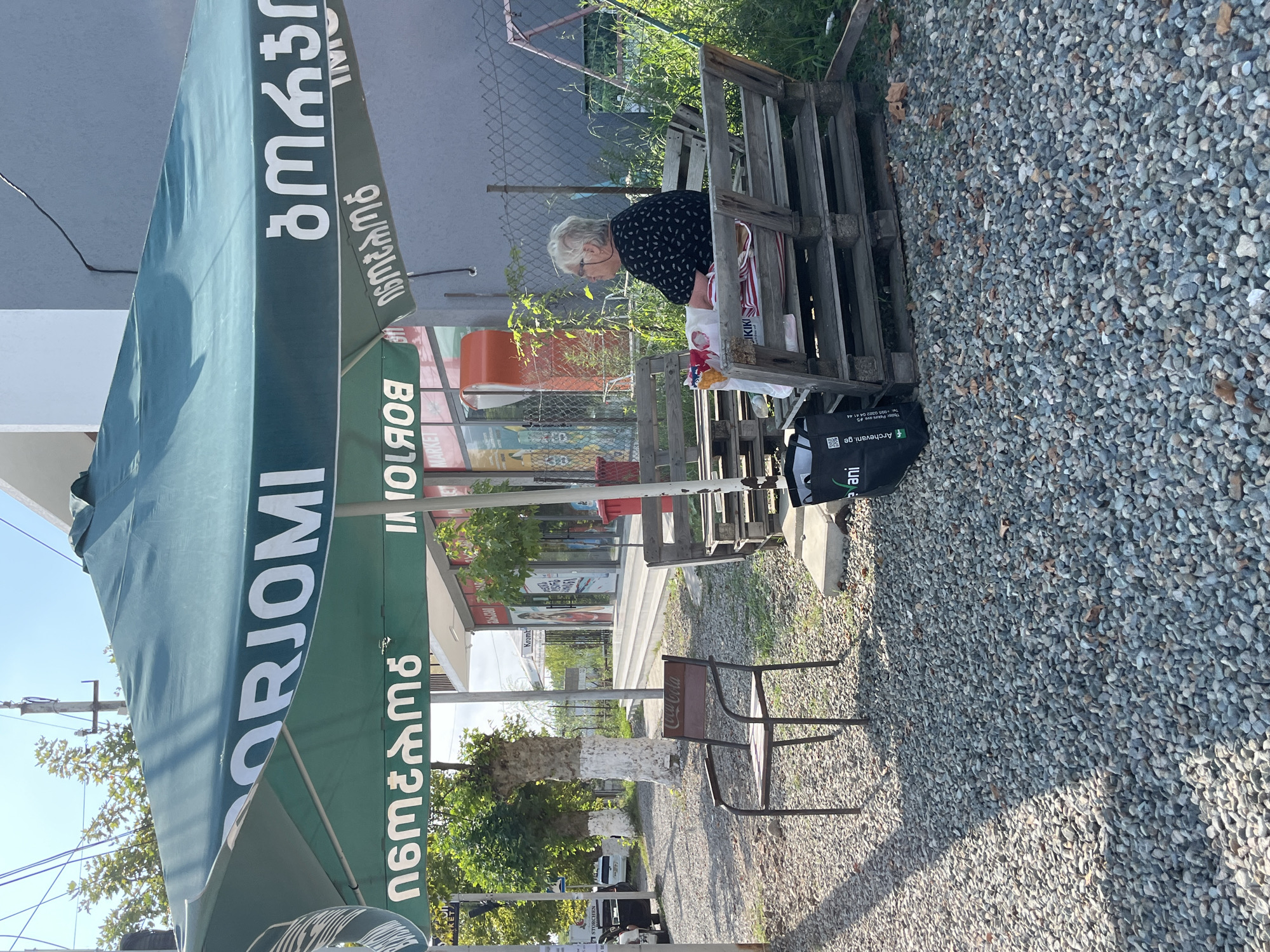 In Ureki, Georgia, a woman sits on a wooden bench crafted from pallets, facing away from the camera and to the right. She has white hair and wears glasses and a dark, patterned shirt. Next to her is a white plastic bag containing food items, and on the bench are striped cushions. The bench is situated outdoors on a gravel ground, shaded by a large green umbrella with white text. In front of the bench, further to the left, is a single metal chair with a Coca-Cola logo on its backrest. Behind the woman and bench, a fence encloses an area with some greenery and a red swing set. To the left, a paved road is visible with parked cars, and a building with storefronts can be seen in the background. The overall scene depicts a roadside seating area, likely outside a shop or cafe, in Ureki.