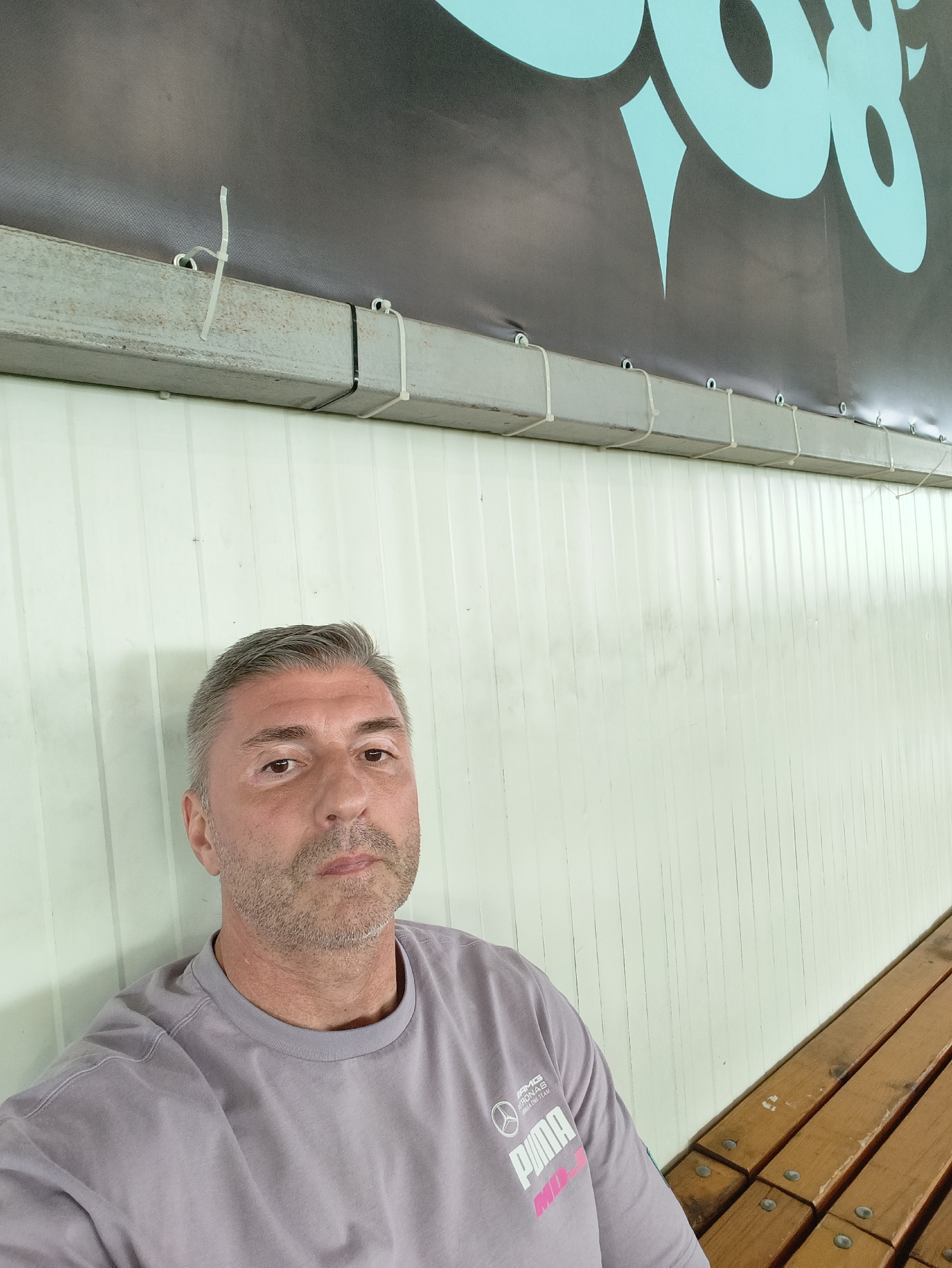 The image shows a middle-aged man with graying hair and stubble, wearing a light purple t-shirt with a Puma logo. He is seated, looking directly at the camera. Behind him is a white corrugated wall and above that, a metal beam secured with zip ties to a dark banner with light blue lettering. To the right, a wooden bench is visible. The scene appears to be indoors or under a covered area, possibly a sports facility or exhibition space. There is no visible indication of the location being in Sofia, Bulgaria.
