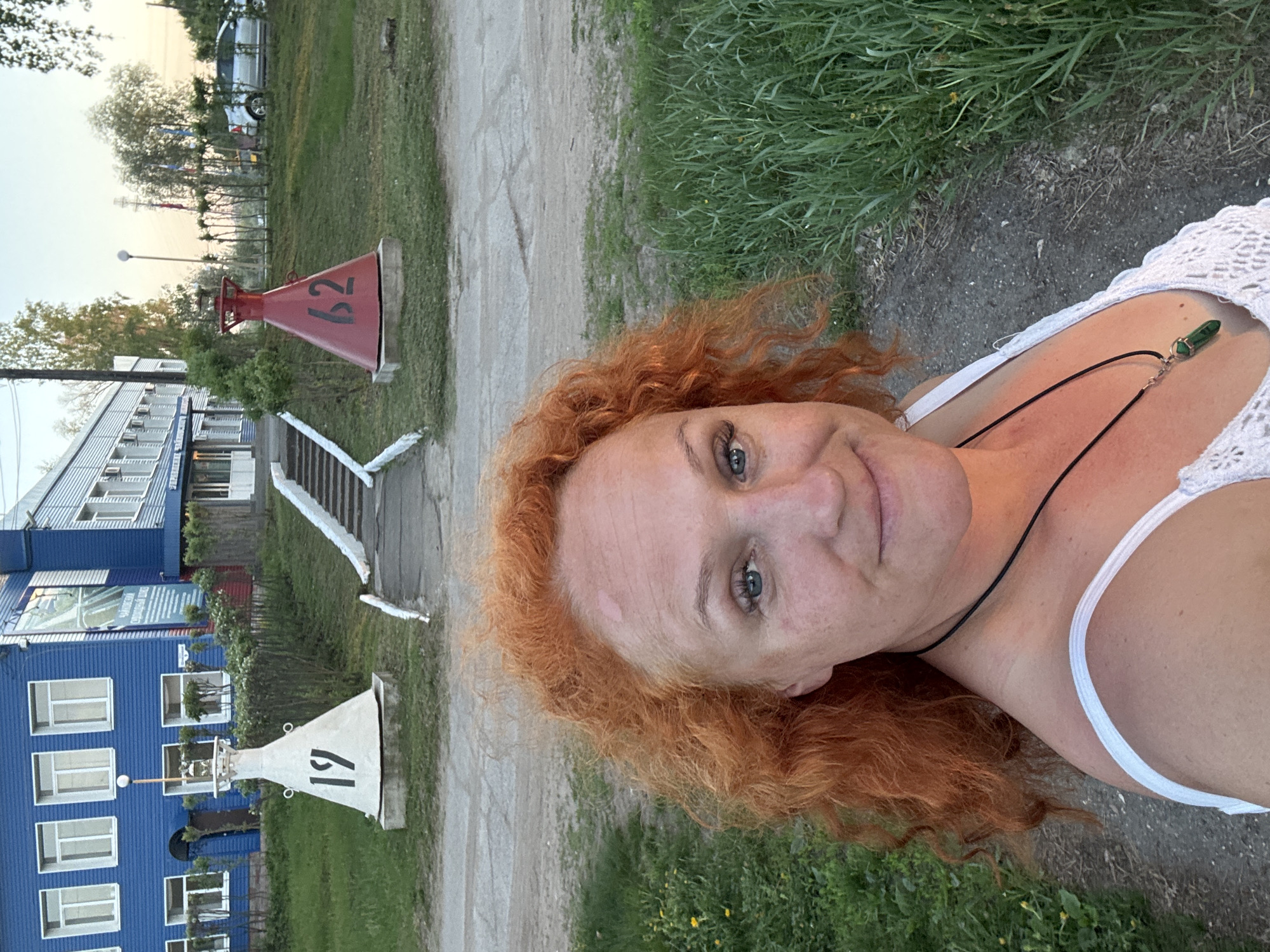 The image shows a selfie of a woman with red curly hair, a white top, and a necklace. In the background are structures and objects in the city of Chaikovsky, Russia. There's a blue multi-story building with white window frames. In front of the building are two navigational buoys with numbers 19 and 62. Between the buoys and the building is a set of white stairs. Further in the background is a car and some trees against a light sky. The woman is positioned in a way that her face is prominently displayed, with grass and a dirt path visible near her.