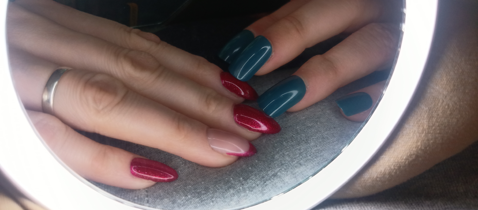Two hands are positioned in front of a lit round mirror, displaying manicured nails. The right hand features a silver ring on the ring finger and displays nails painted in a dark teal color. The left hand shows nails painted in a red, glittery color, with one nail featuring a pale pink base and a red, glittery tip in a French manicure style. The background shows a gray fabric-like surface. The scene appears to be indoors, possibly in a home setting, but the specific location is unknown.